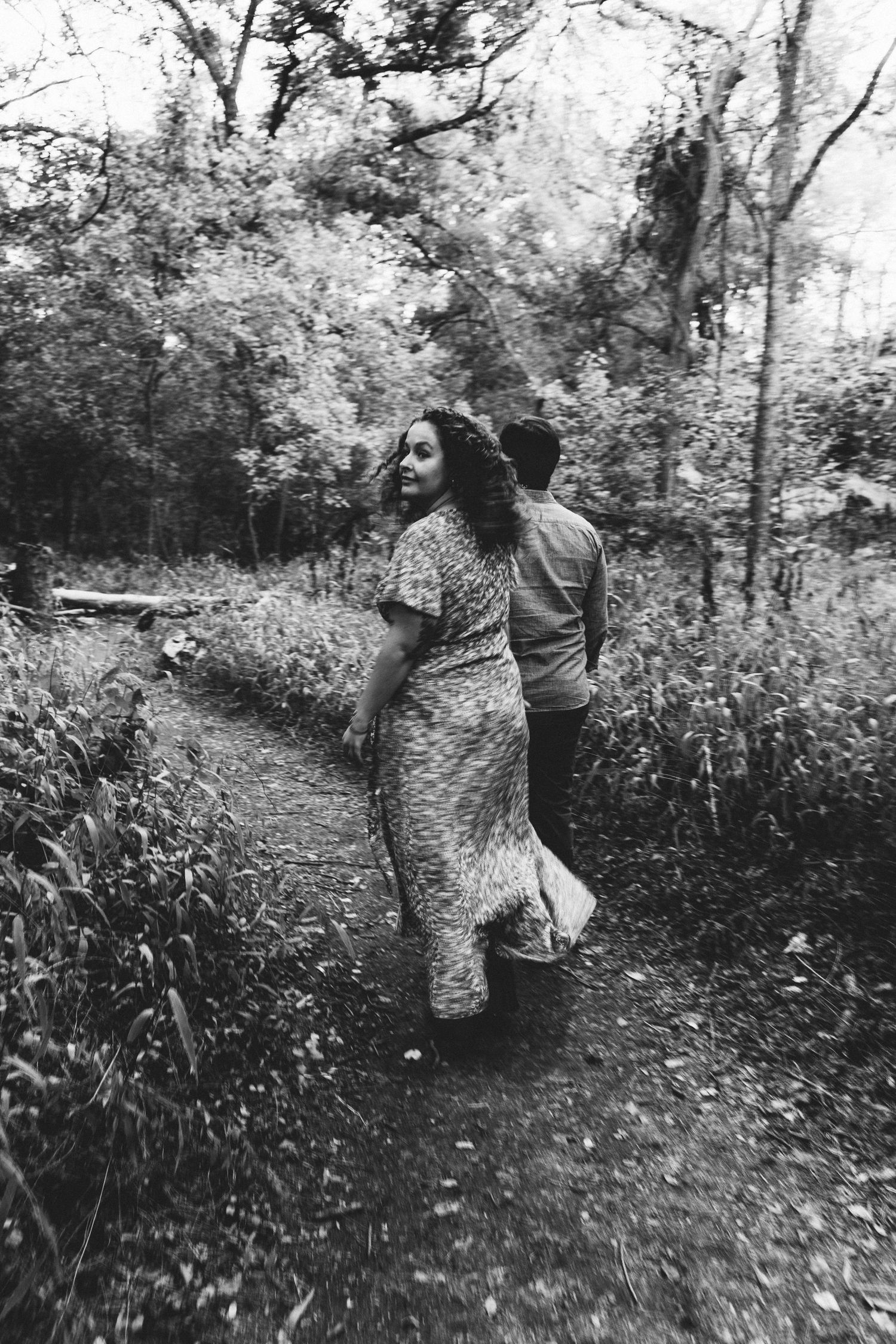 Black and white photo of couple walking along a narrow forest trail, woman in floral dress.