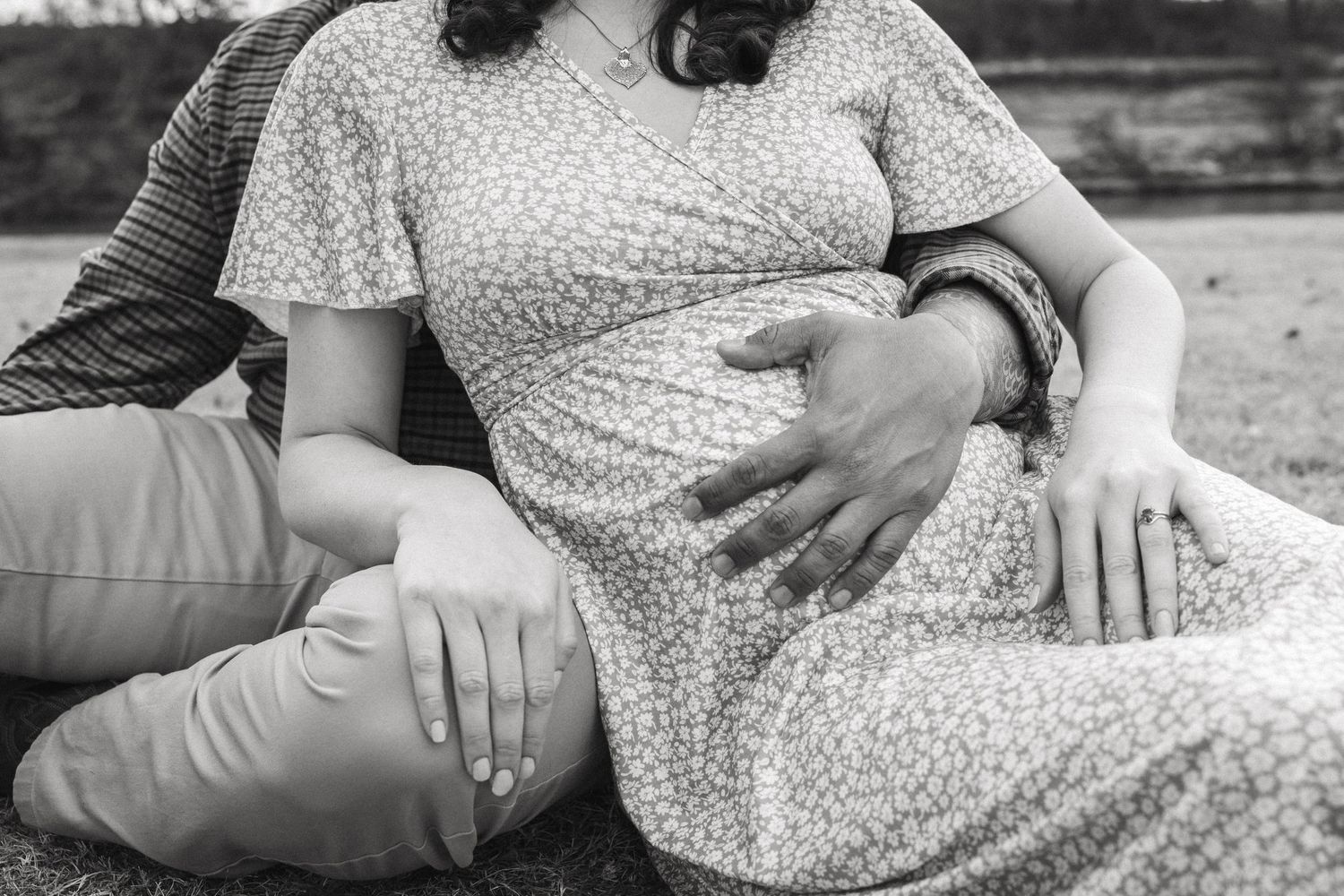 Black and white close-up of a pregnant woman in a floral dress cradling her baby bump while sitting outdoors.