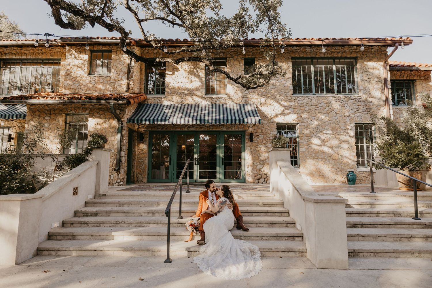 Wedding couple sitting on steps outside a rustic stone venue building with arched green doors and sprawling oak trees.