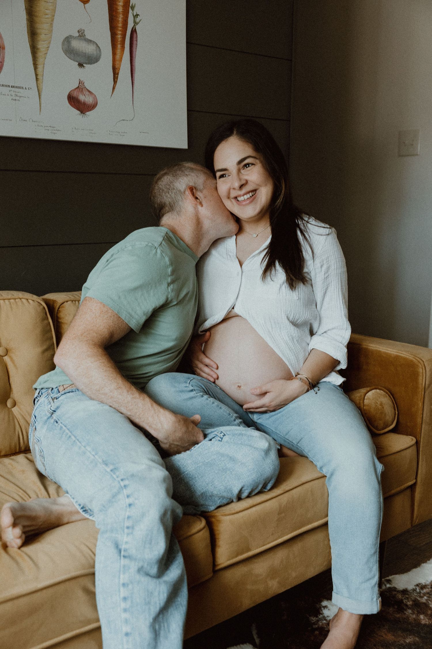 A smiling pregnant woman receives a gentle kiss from her partner while sitting on a yellow sofa.