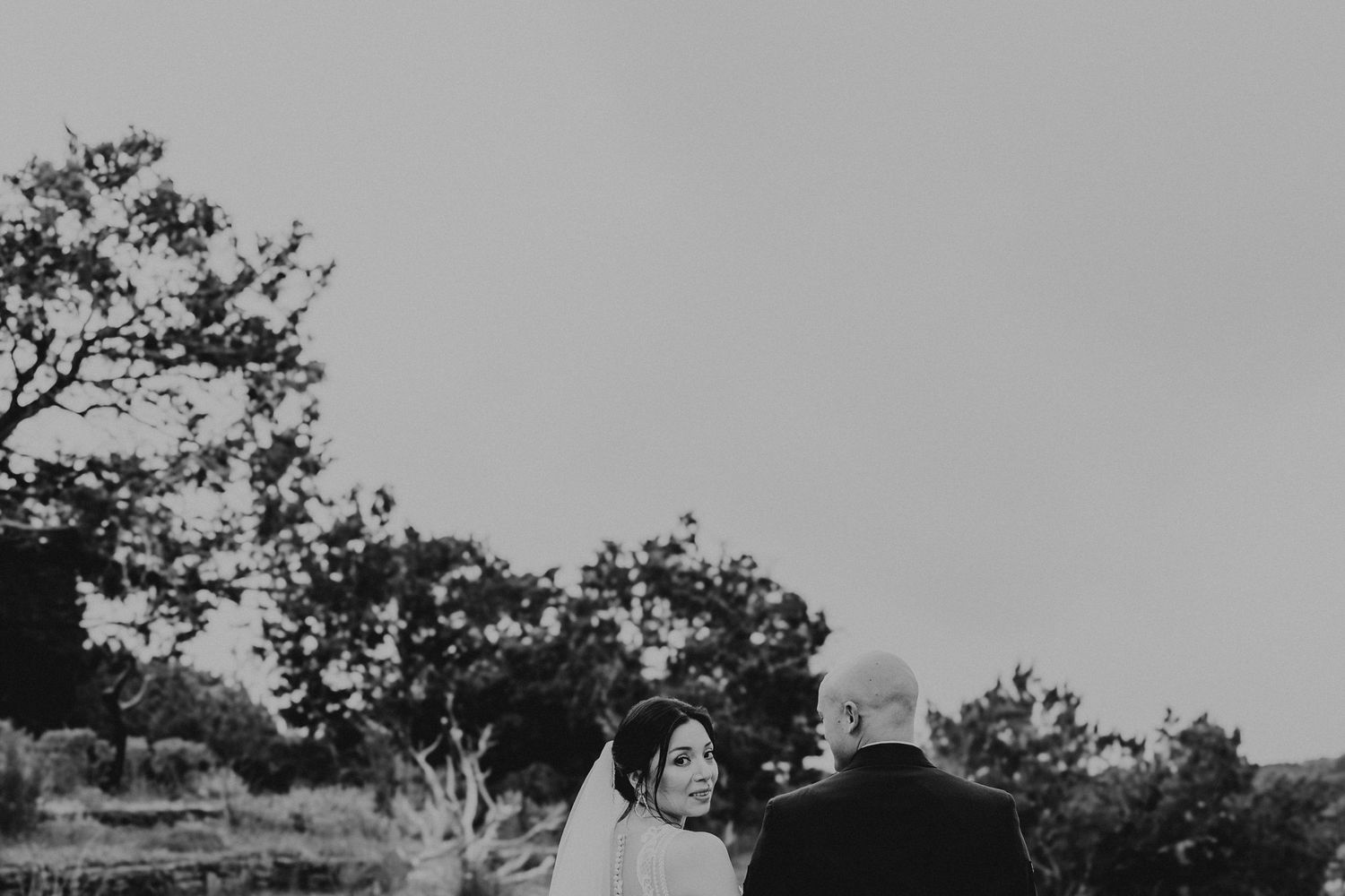 Black and white photo of a bride in a veil looking back at her partner outdoors.