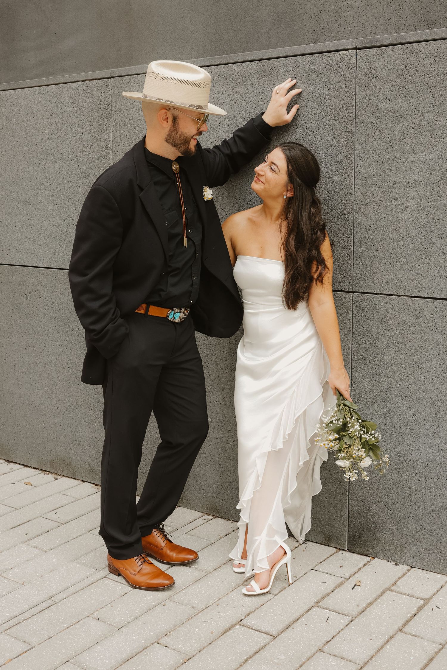 Groom in black suit and cream hat stands with smiling bride in white strapless gown against gray wall.