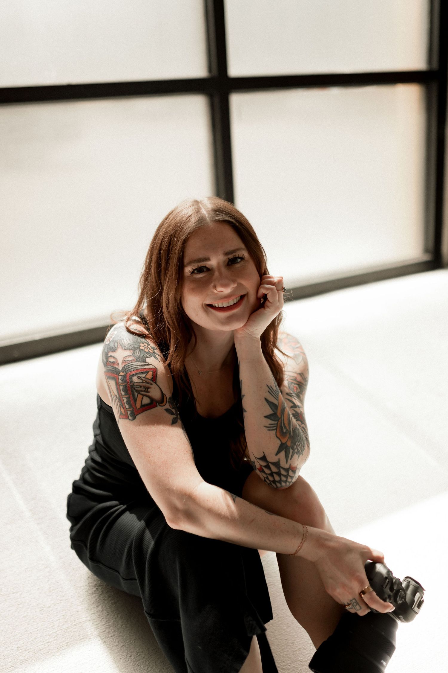 Smiling woman with tattoos on her arms sits casually on the floor in black outfit near a large window with her camera.