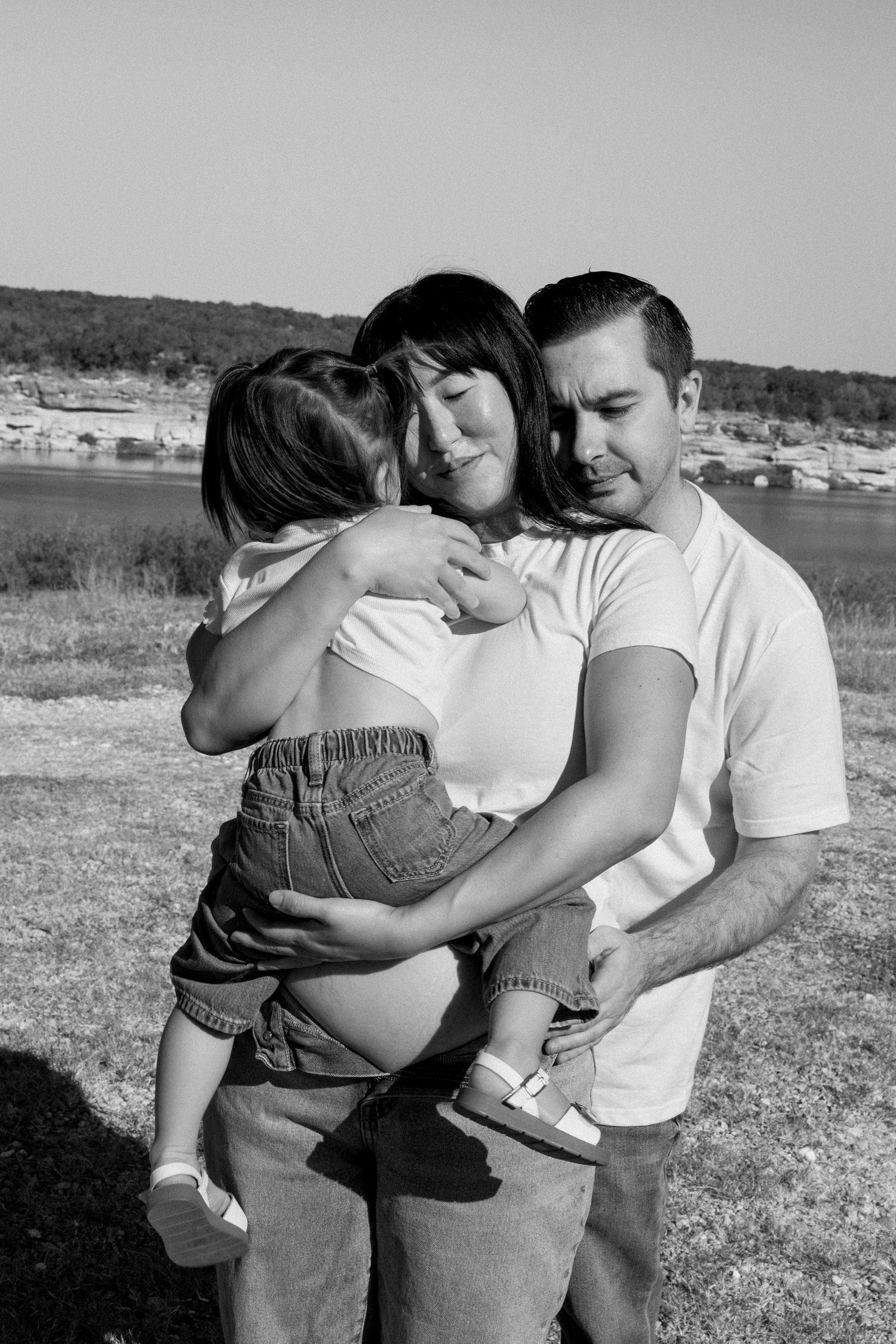 Black and white photo of parents holding their young child together near a lake outdoors.