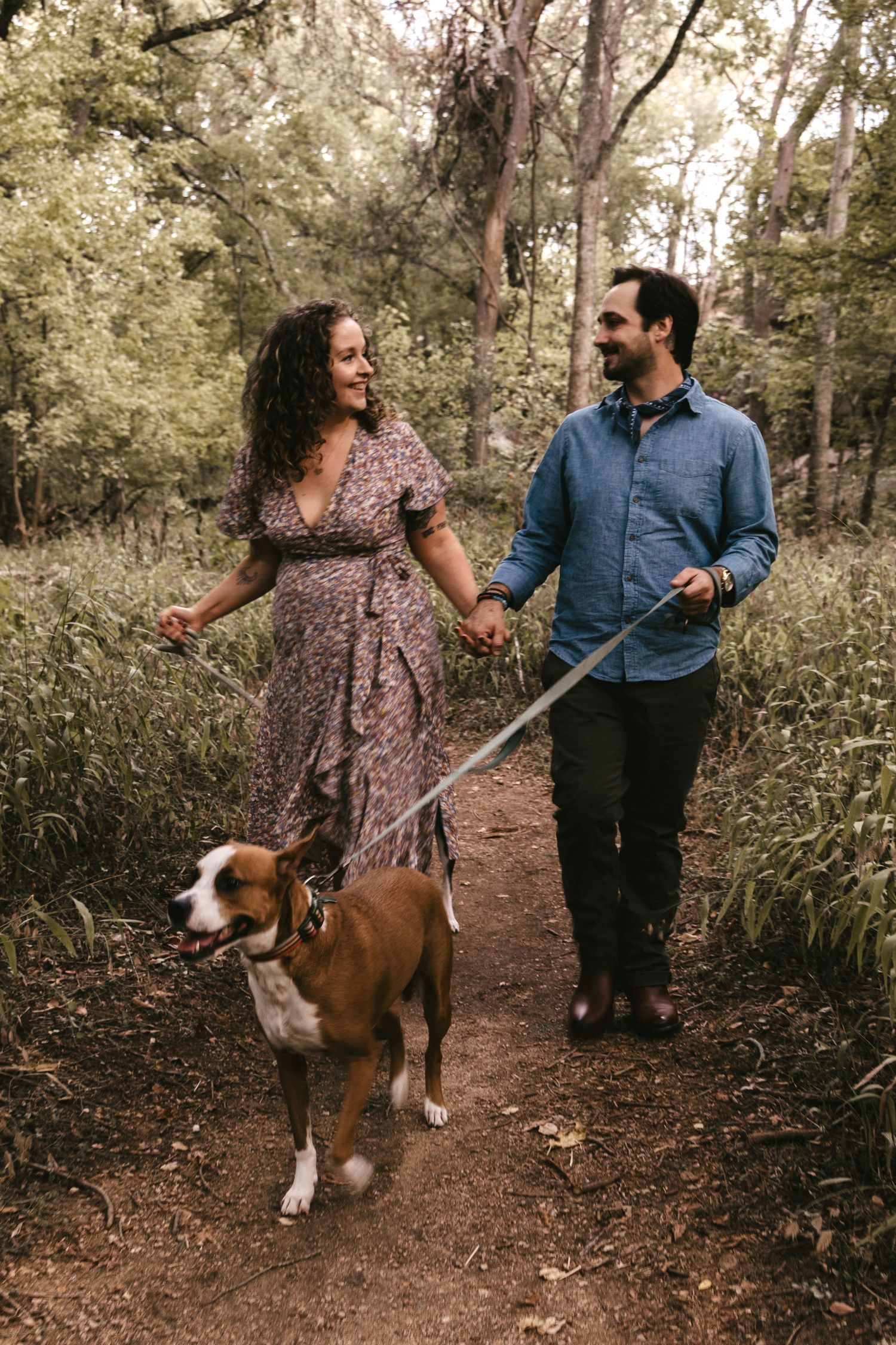 A joyful couple walks hand in hand with their brown and white dog along a lush green forest trail.