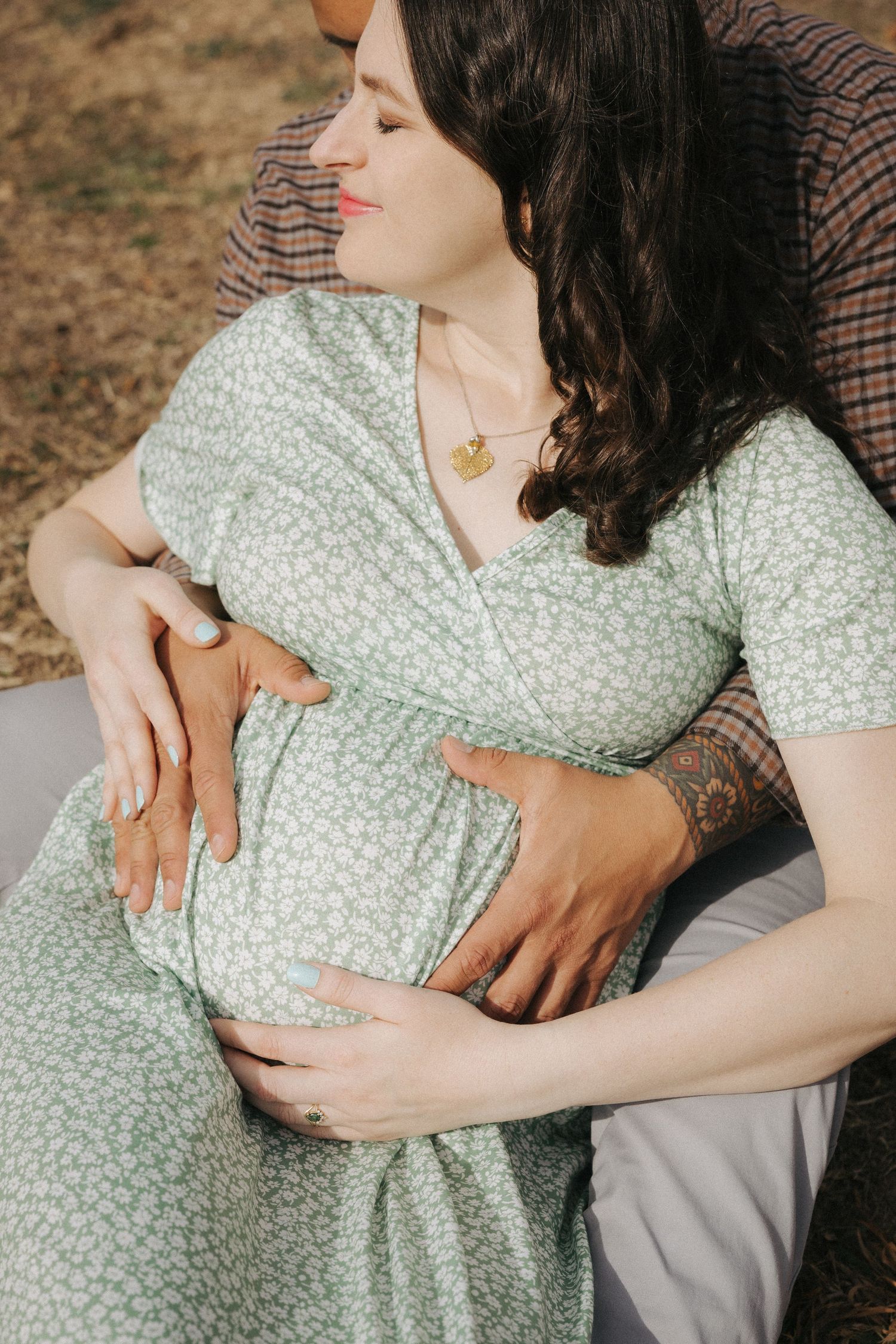 Close-up of pregnant woman in mint floral dress with partner embracing her baby bump outdoors.