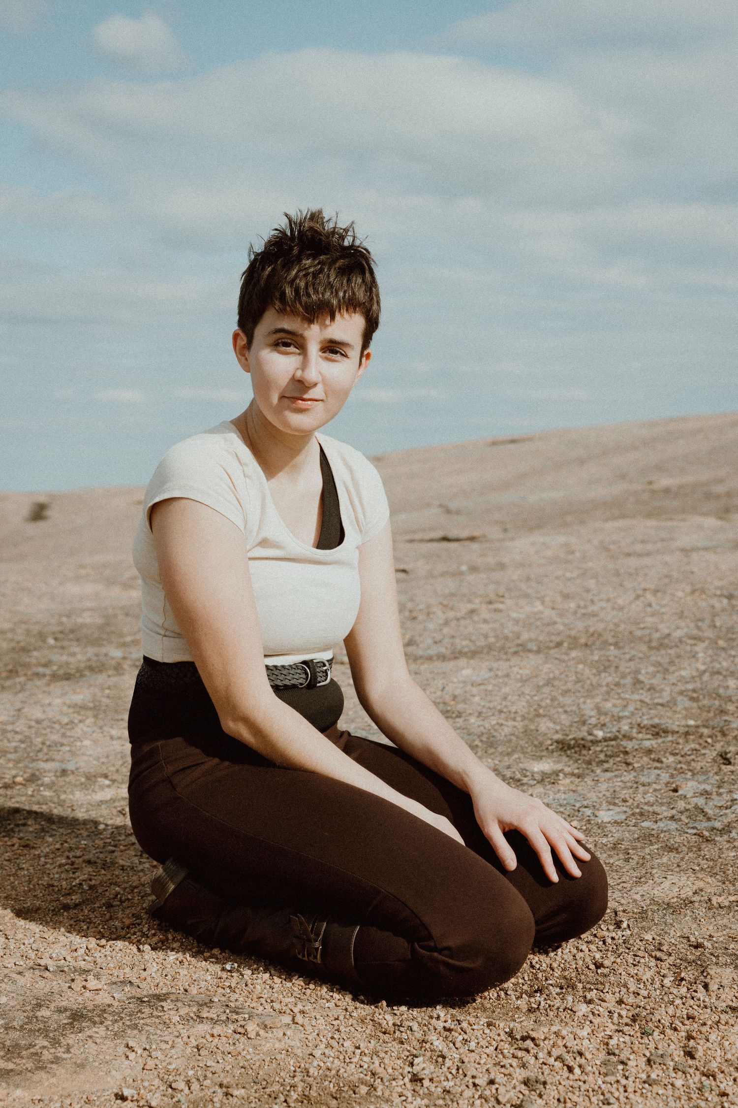 Person with short hair in white top and dark pants sitting on sandy rocky ground.