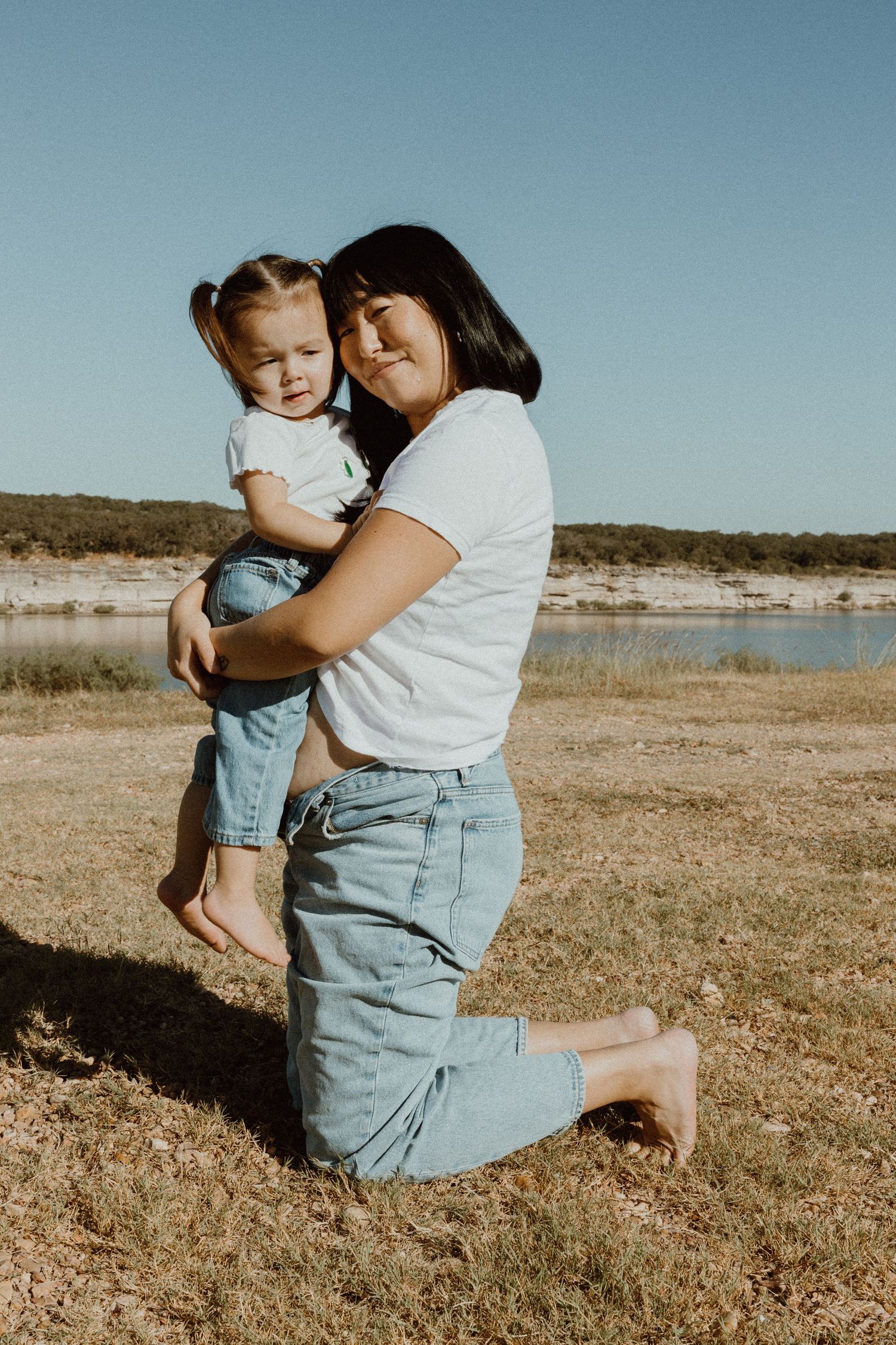 Mother kneeling on grass hugging her toddler daughter near a scenic lake on a sunny day.
