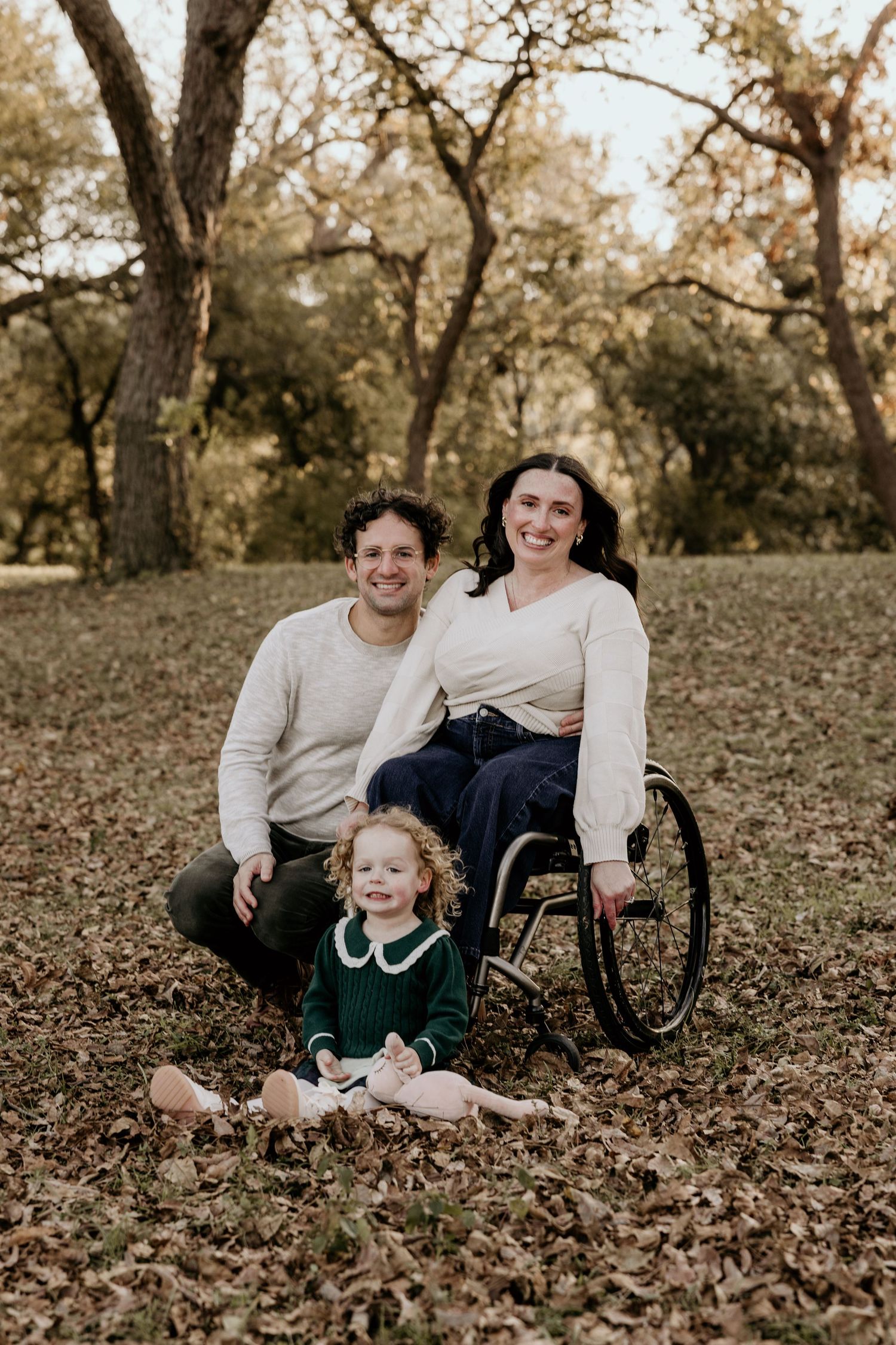 Family portrait with man, woman in wheelchair, and toddler girl in green dress sitting on autumn leaves in park.