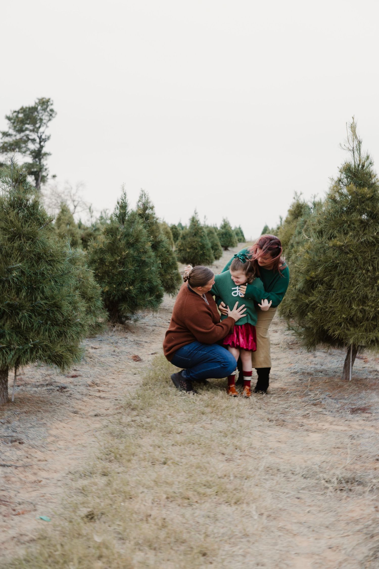 Two mothers crouching down hugging young daughter between rows of Christmas trees outdoors.