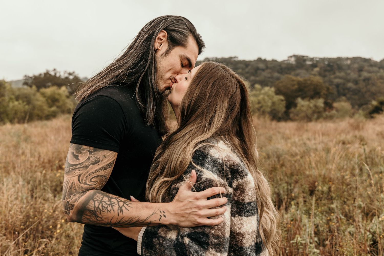 Tattooed man with long hair kissing his girlfriend in an autumn field.