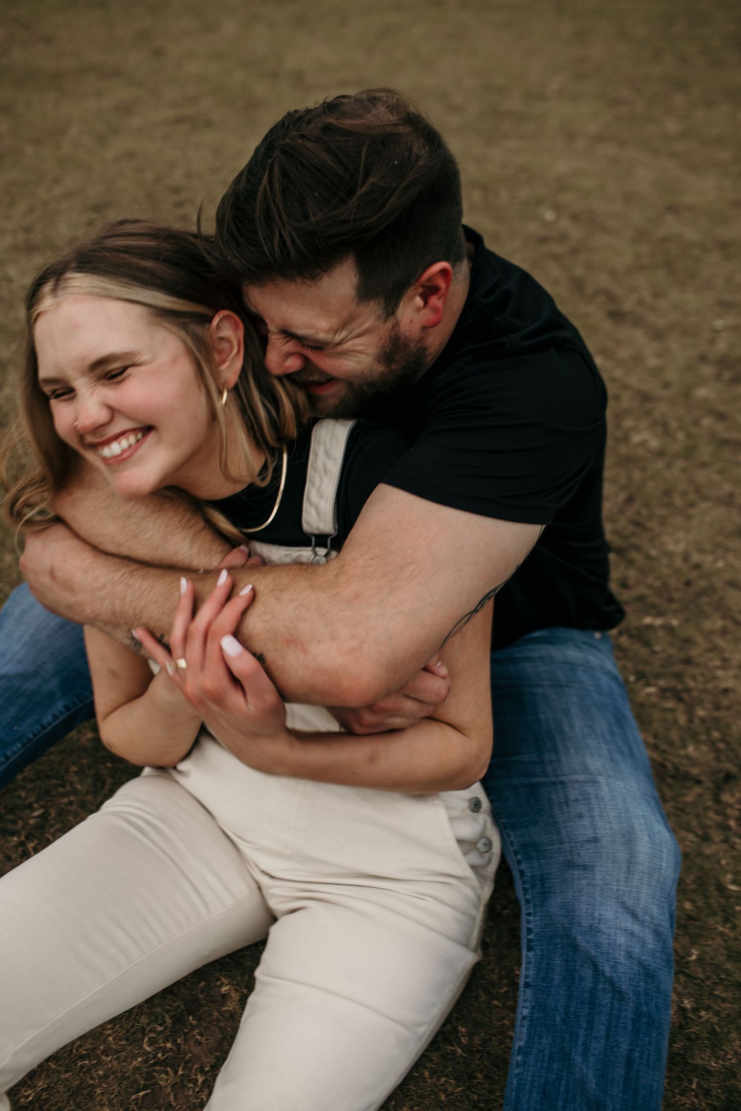 Couple sitting on grass laughing joyfully, man hugging woman from behind in a candid engagement photo.