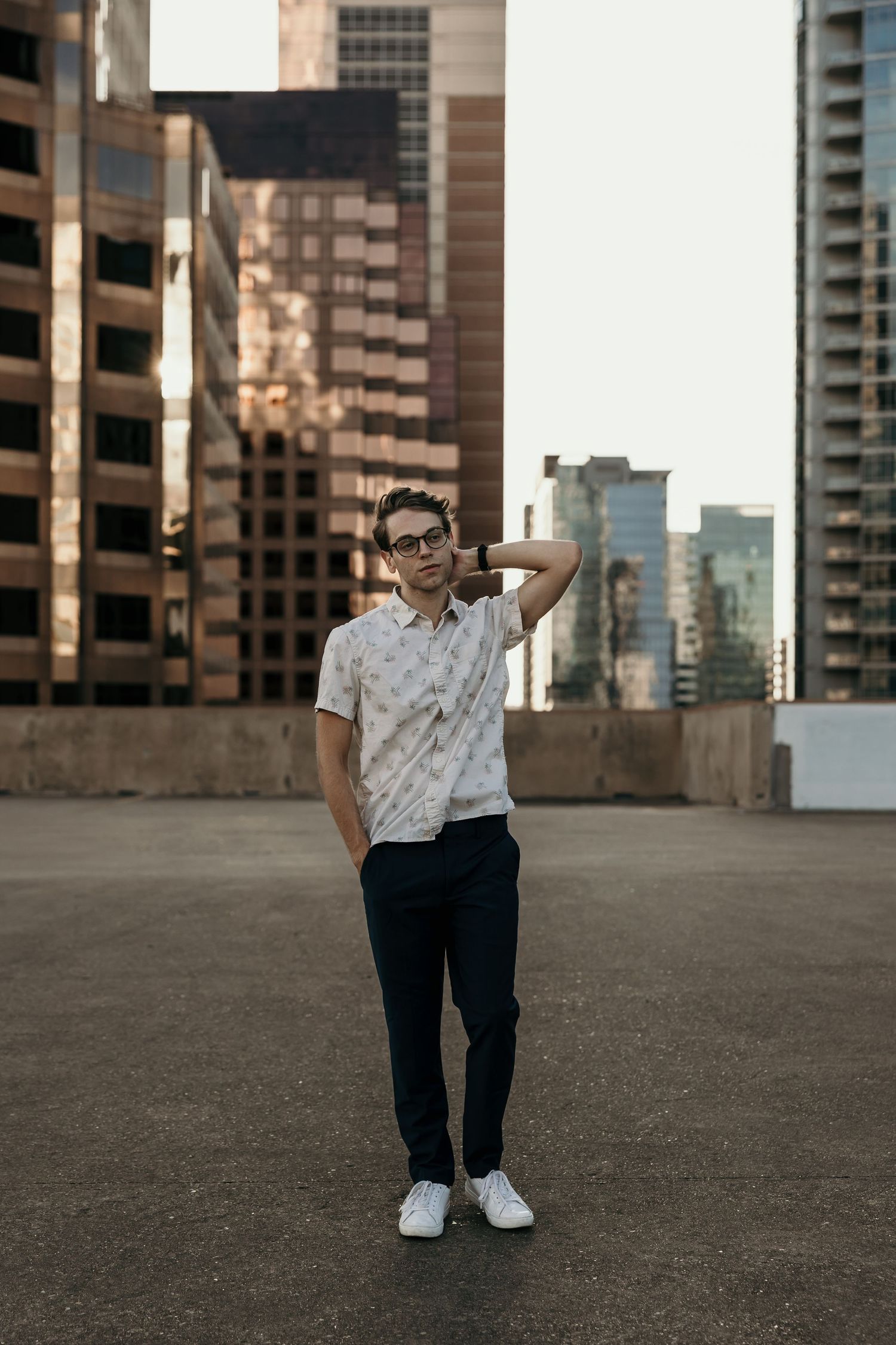 Young man in patterned shirt and glasses posing on urban rooftop with city skyline.
