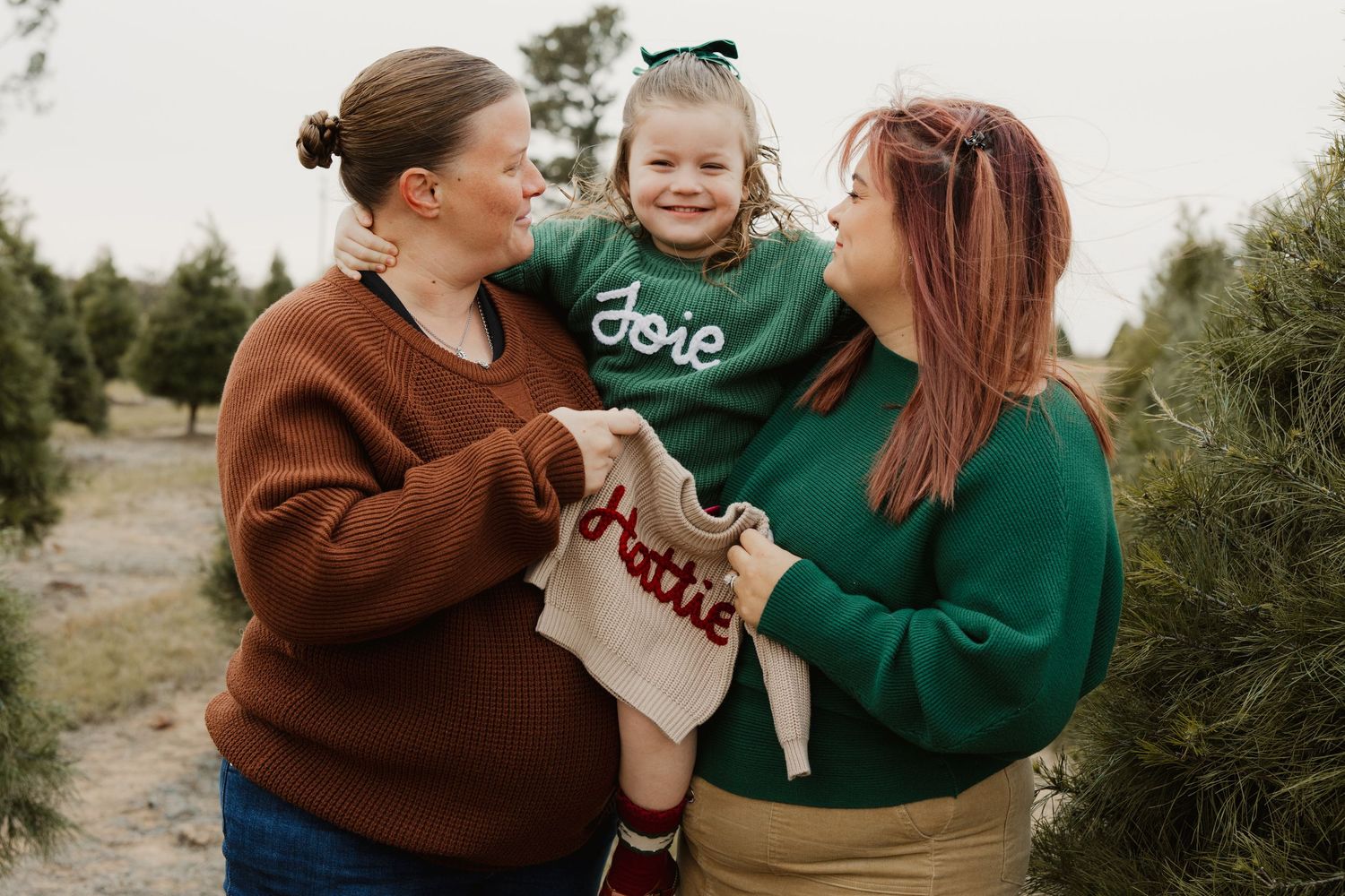 Two women holding smiling toddler girl in green sweater at outdoor Christmas tree farm.