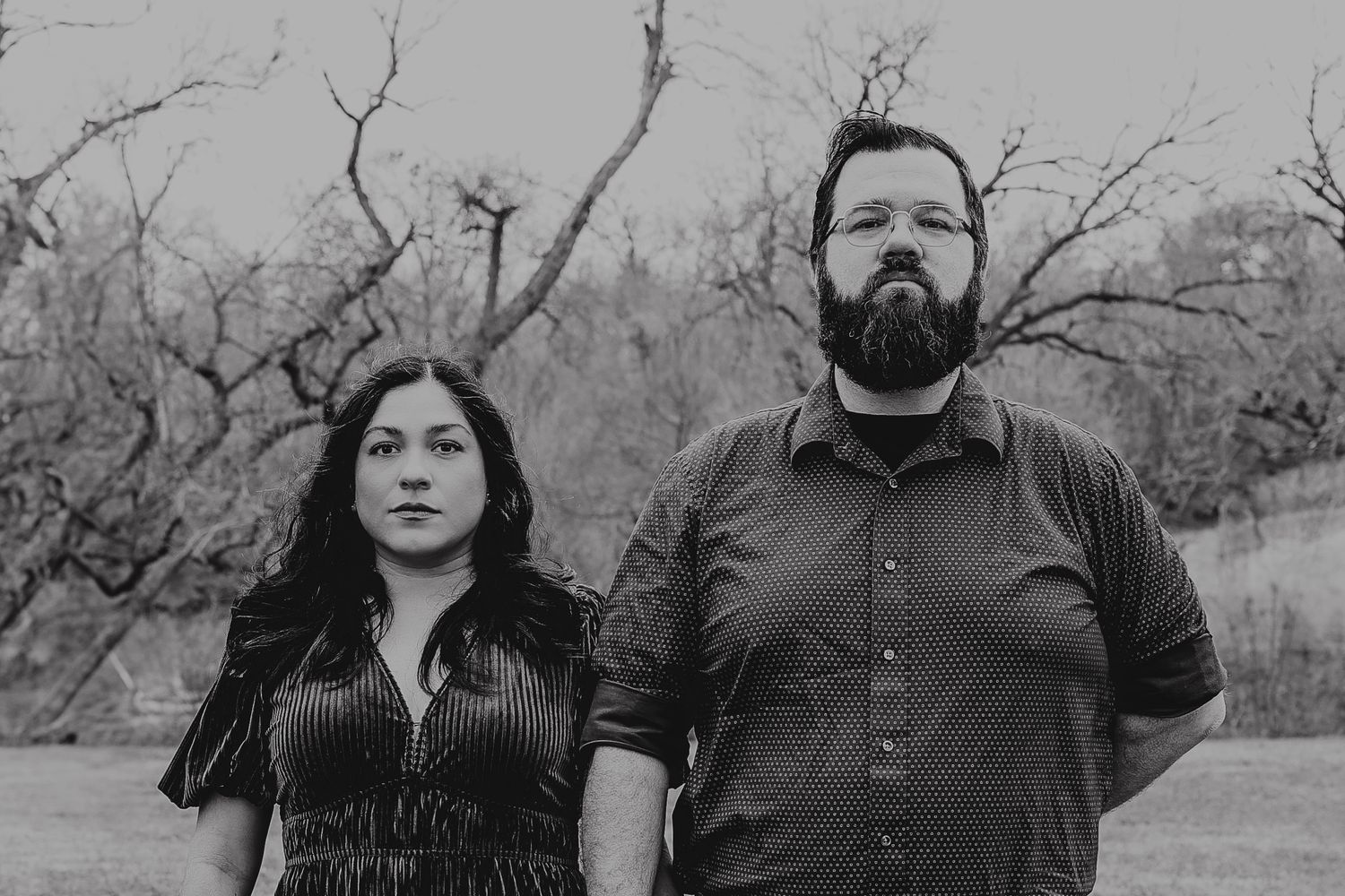 Black and white portrait of a couple standing together outdoors with bare trees behind them.