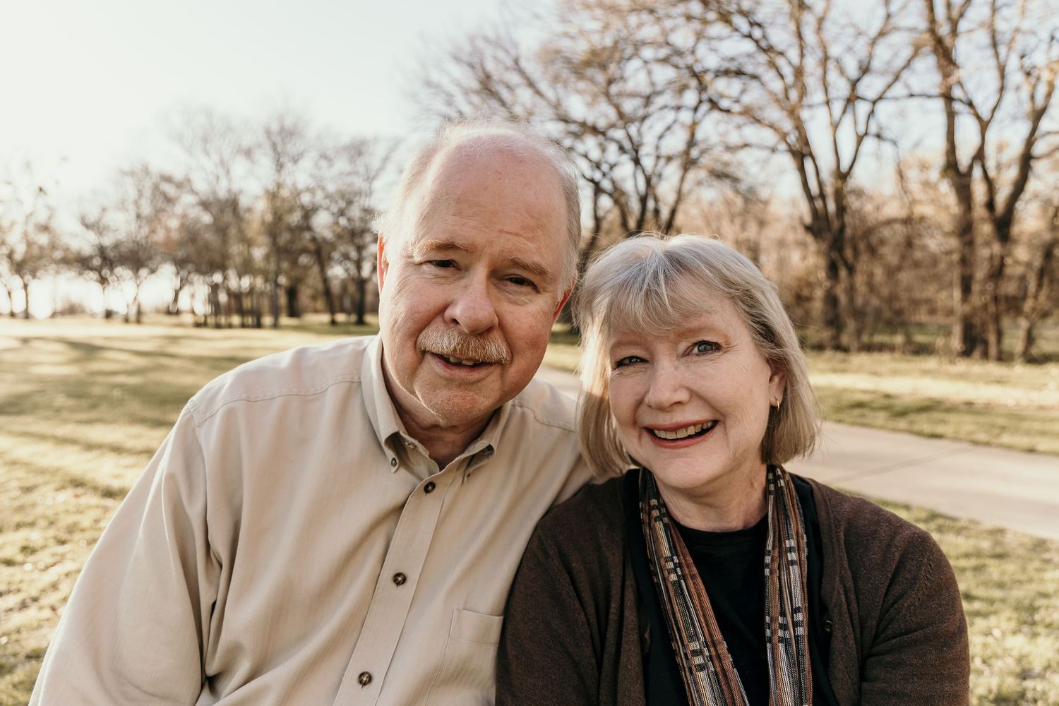Close-up portrait of a smiling older couple outdoors with a sunny park background.