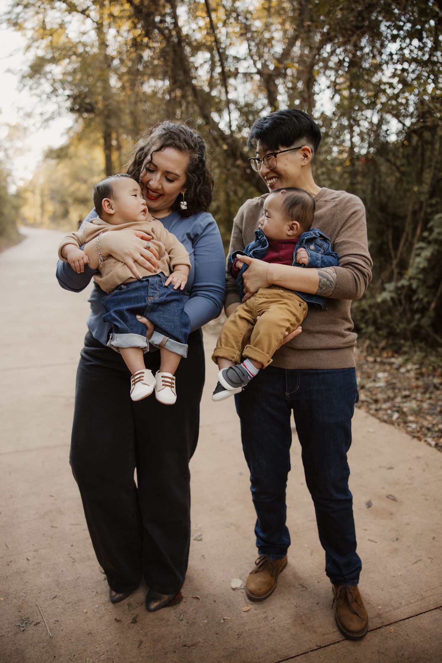 Happy family of two parents each holding an infant on a scenic tree-lined outdoor pathway.