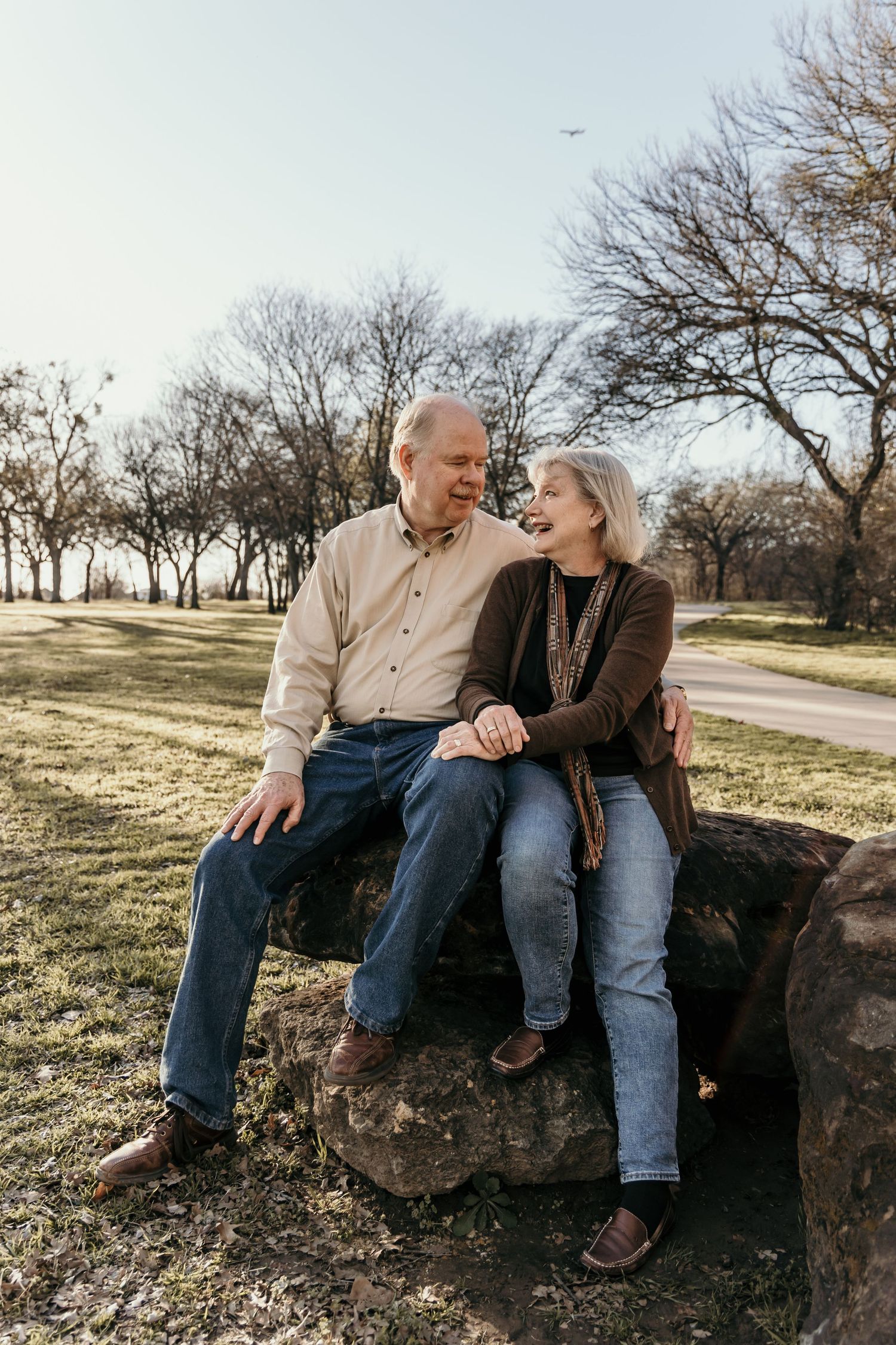 Older couple sitting on rocks in a sunny park, smiling at each other with bare trees behind.