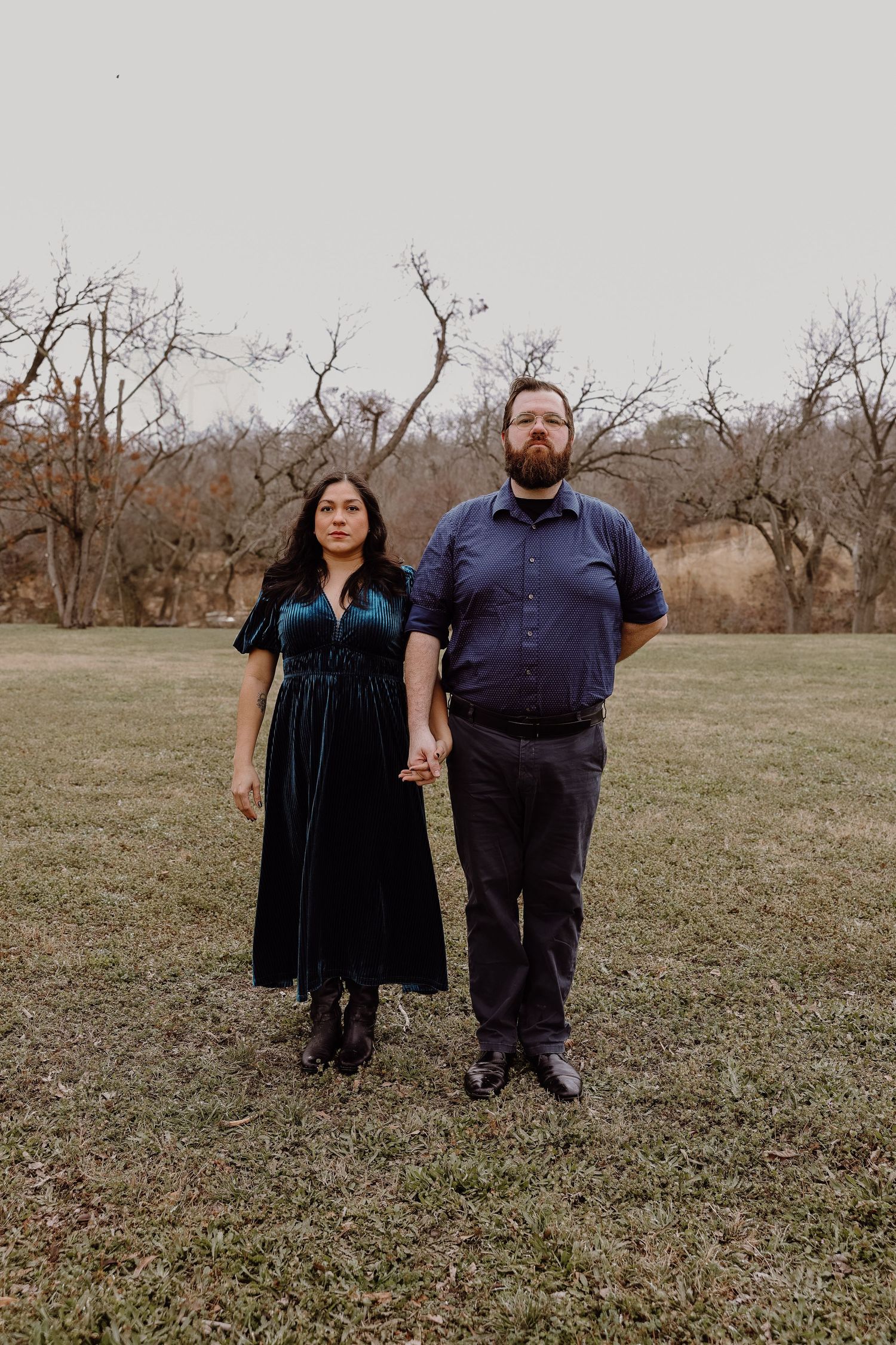 Couple standing hand in hand in a grassy field with bare winter trees in the background.