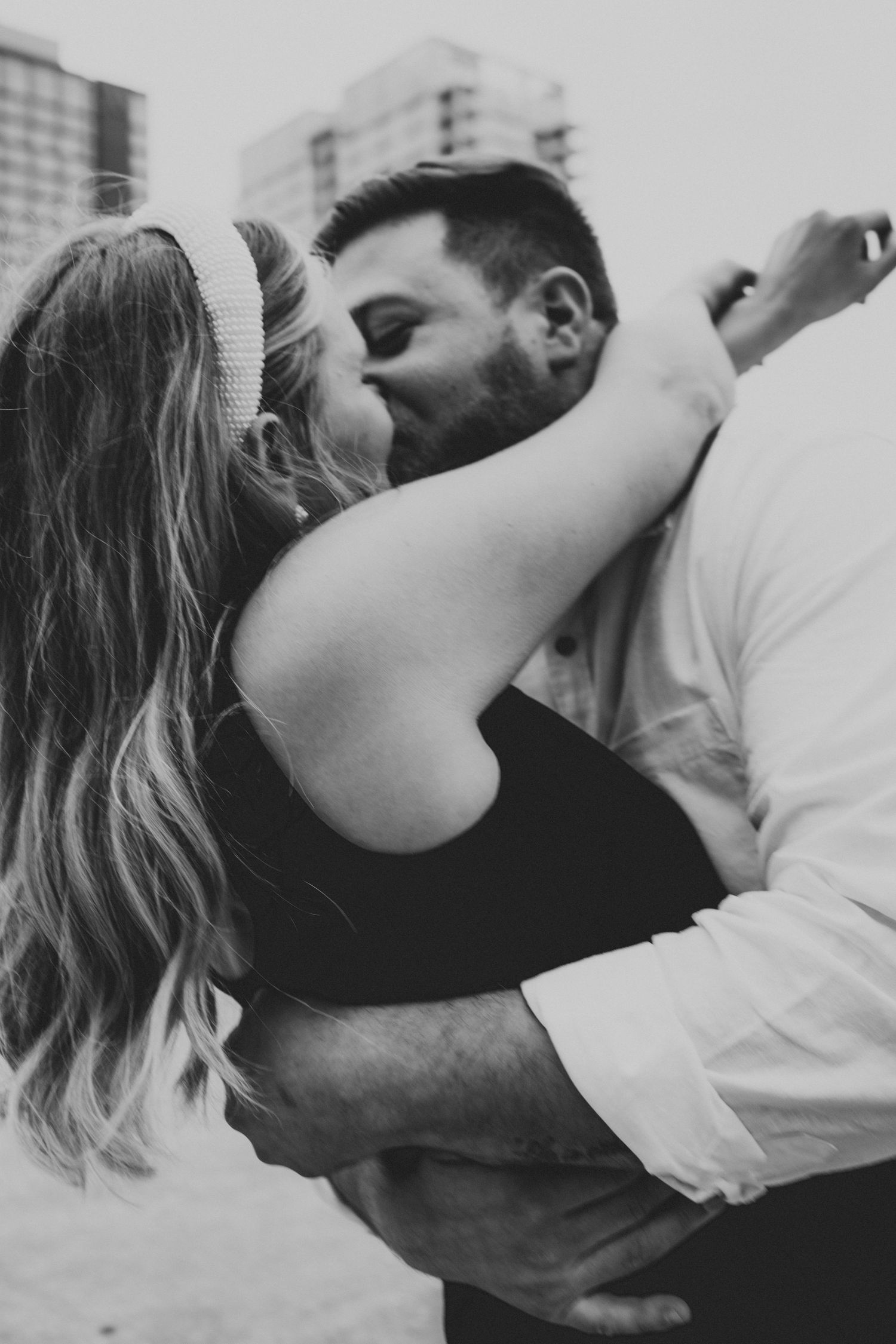 Black and white close-up of couple sharing a passionate kiss outdoors with city skyline backdrop.