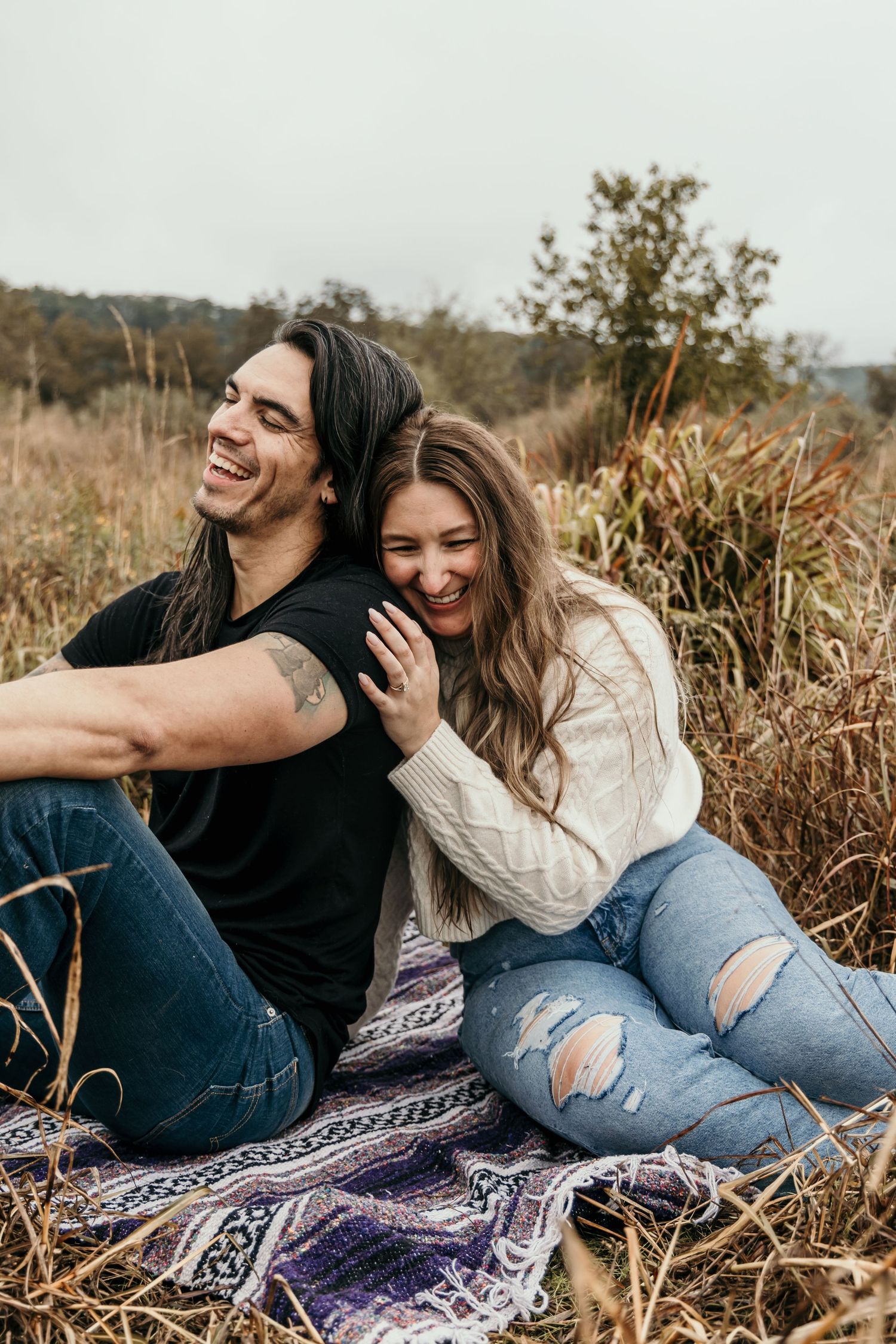 Laughing couple sitting back-to-back on a blanket in an autumn field, enjoying a joyful moment.