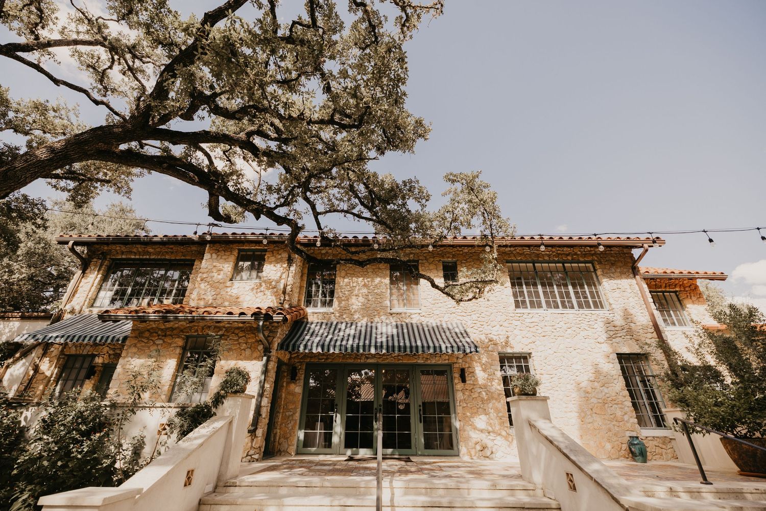 Elegant stone venue with Spanish-style architecture, tiled awning, and string lights beneath large oak trees.