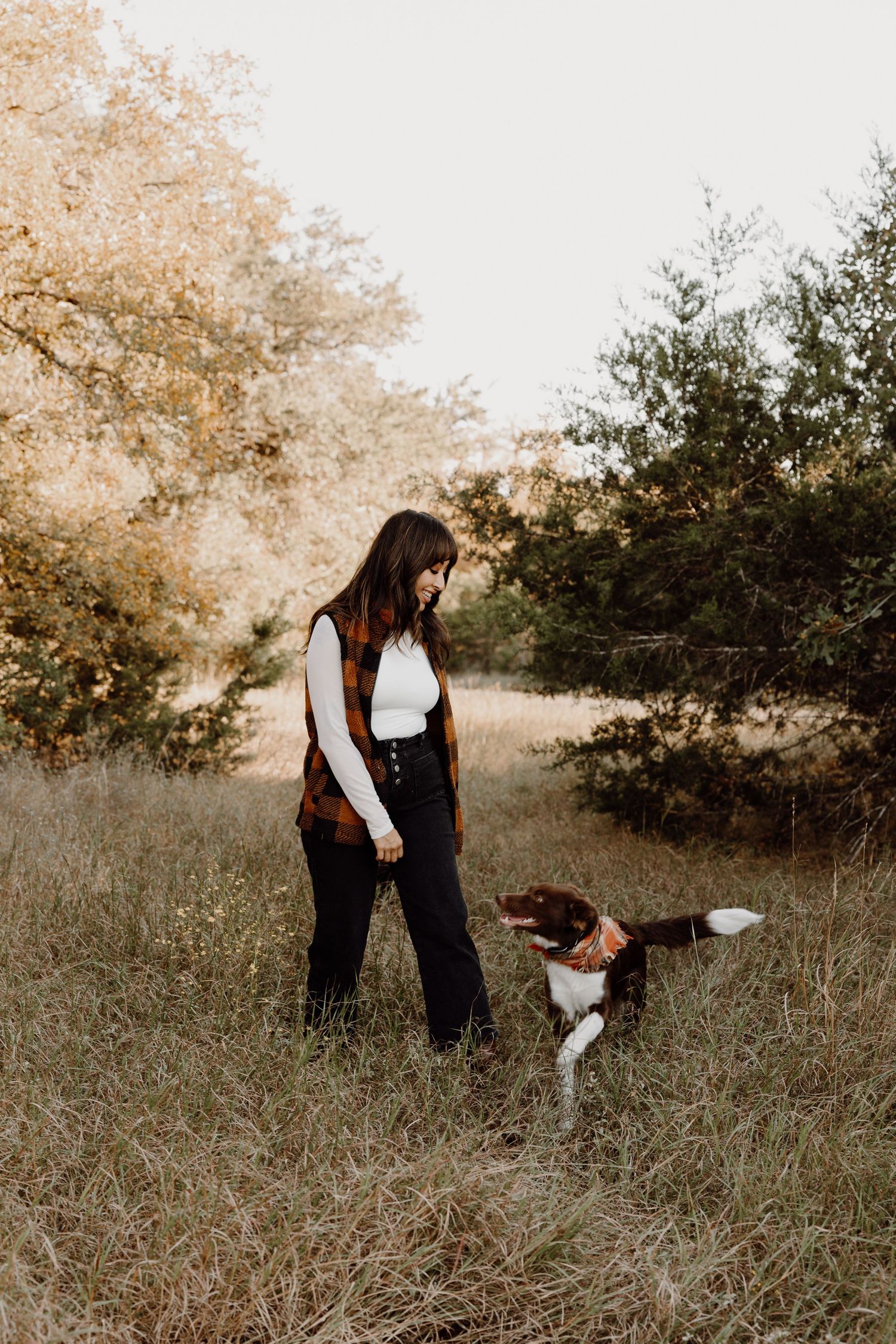 Woman in plaid vest interacting with a brown and white dog in a scenic grassy meadow.