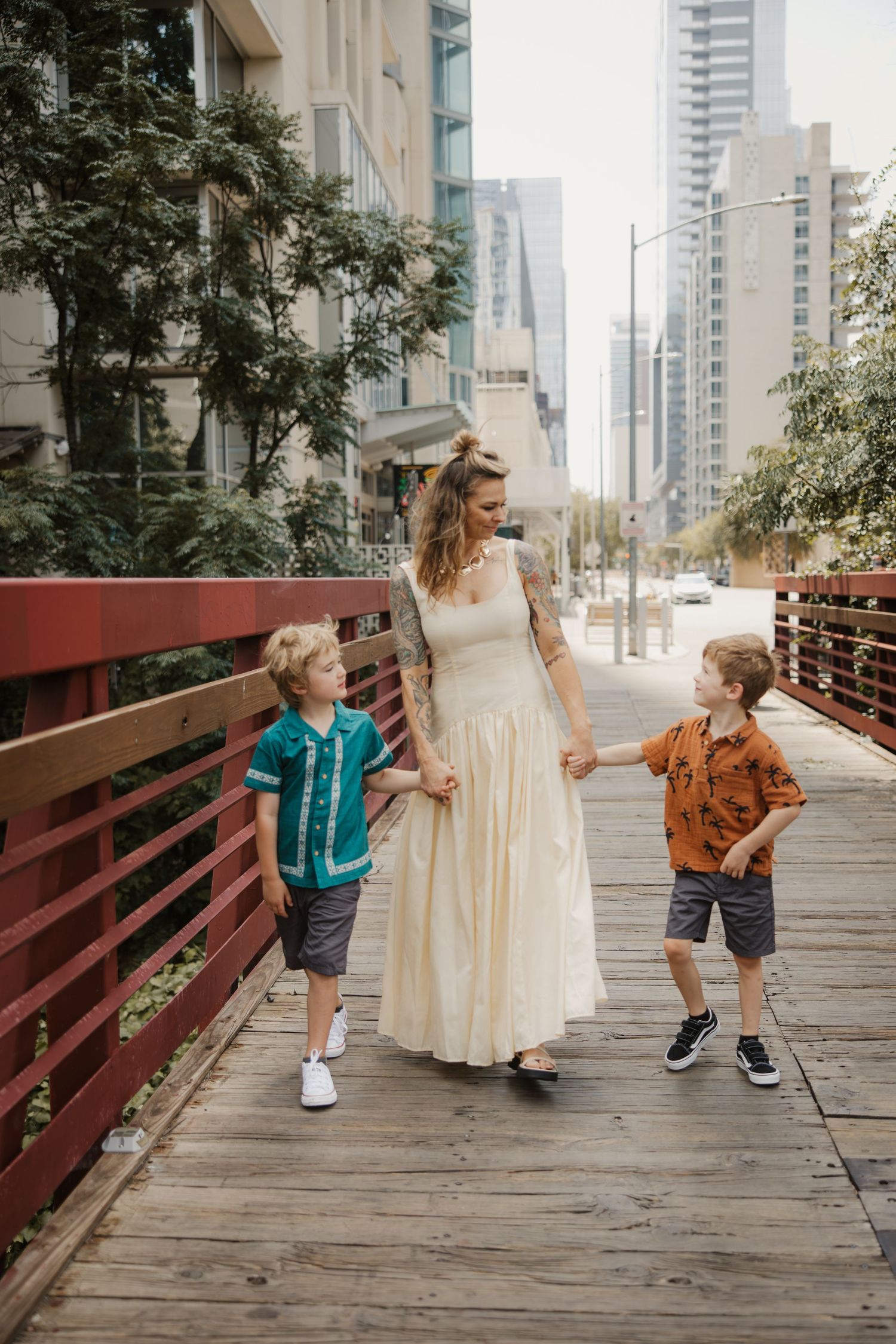 A mother in a flowing cream dress walks hand-in-hand with her two young sons on a city bridge.