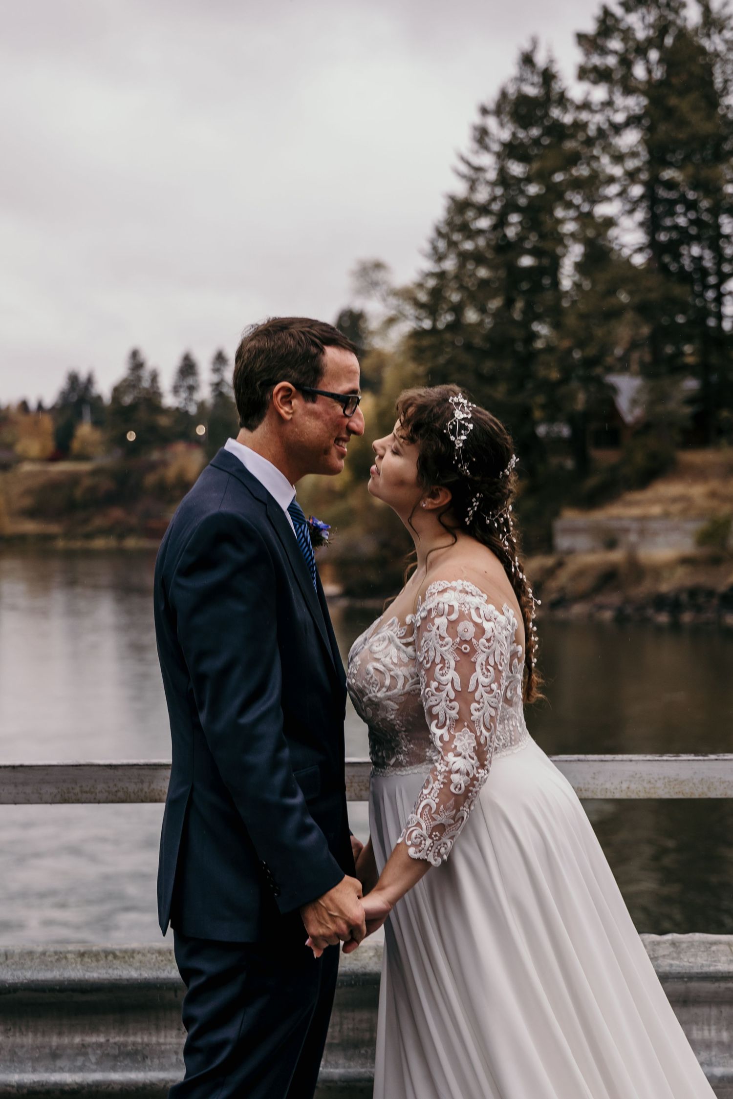 Bride in lace gown and groom in navy suit hold hands and gaze at each other by a lakeside bridge.