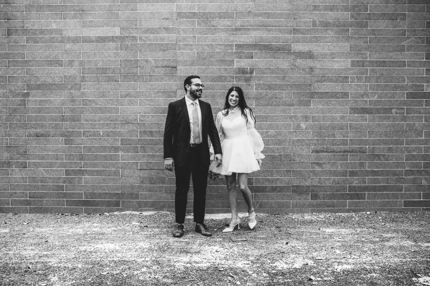Black and white photo of bride and groom holding hands and smiling in front of a brick wall.