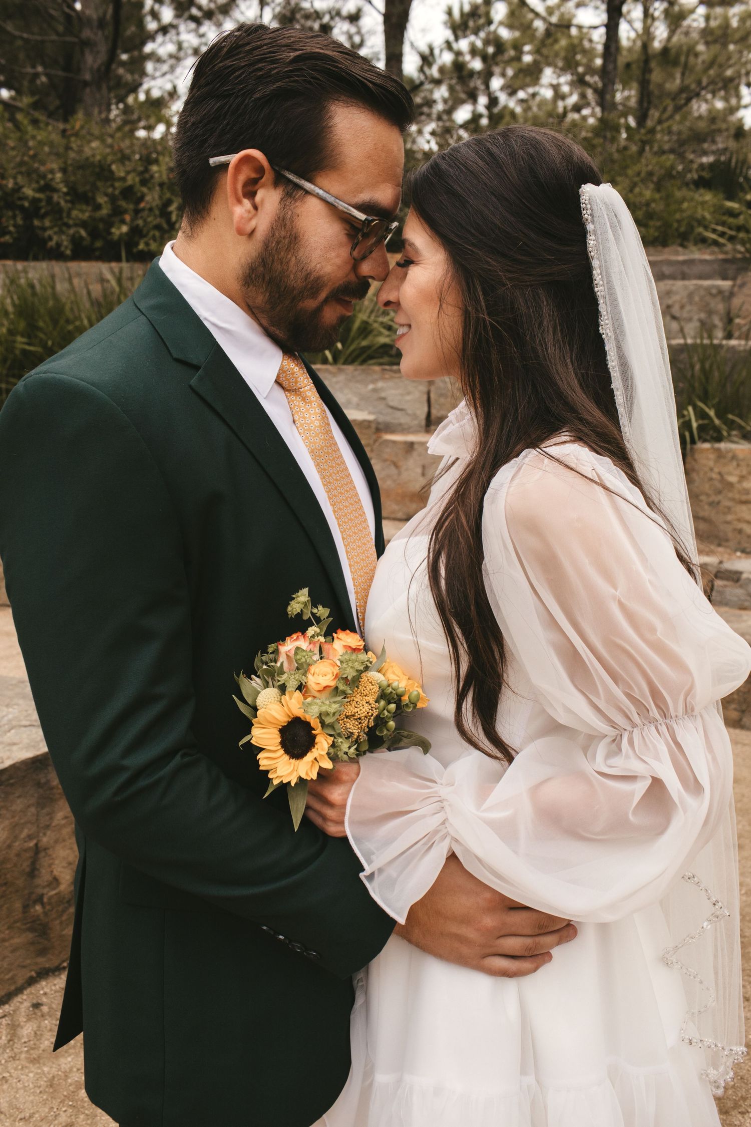 Bride and groom touch foreheads romantically while holding sunflower bouquet in outdoor wedding portrait.
