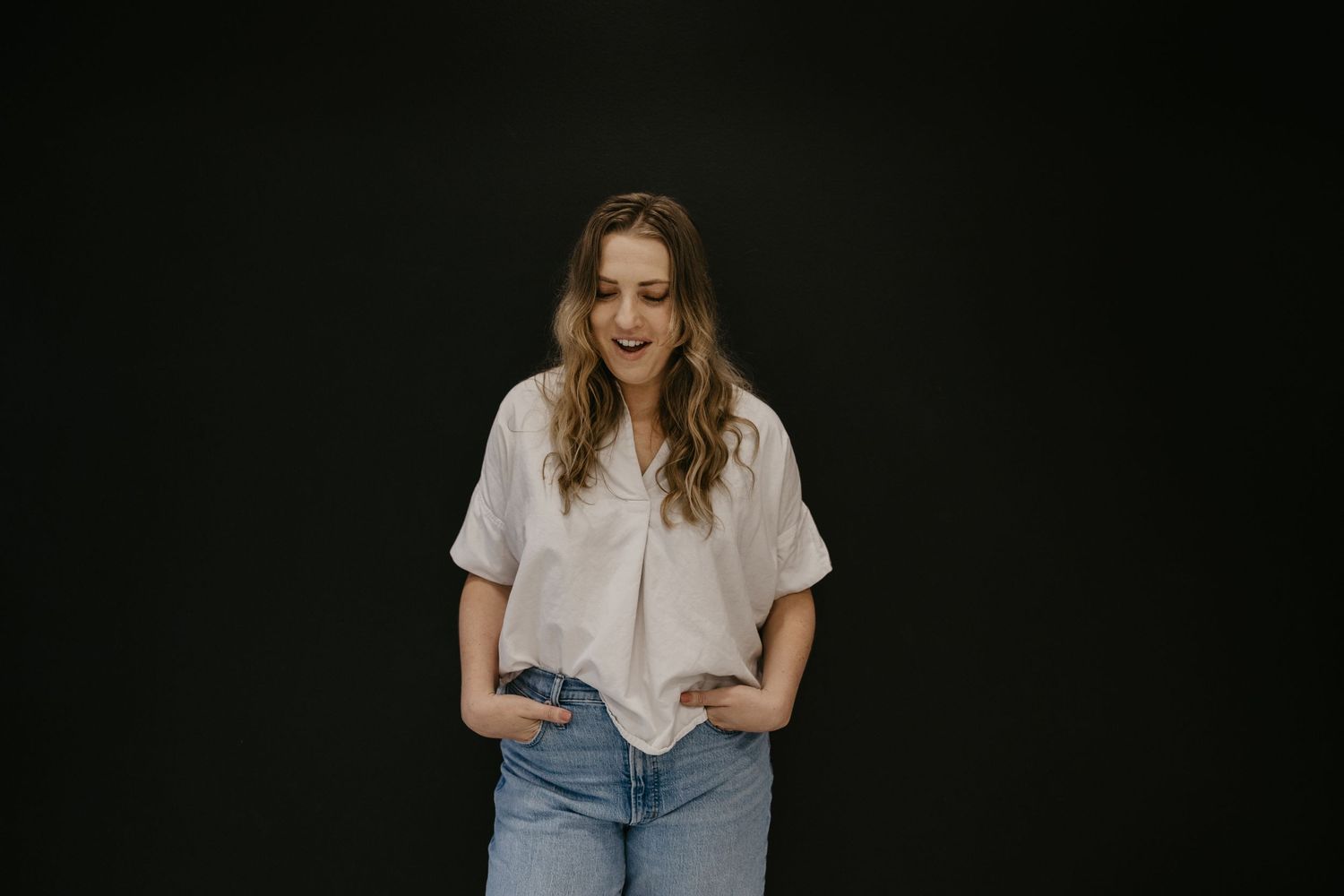 Smiling young woman in white blouse and light jeans posing against a dark background.