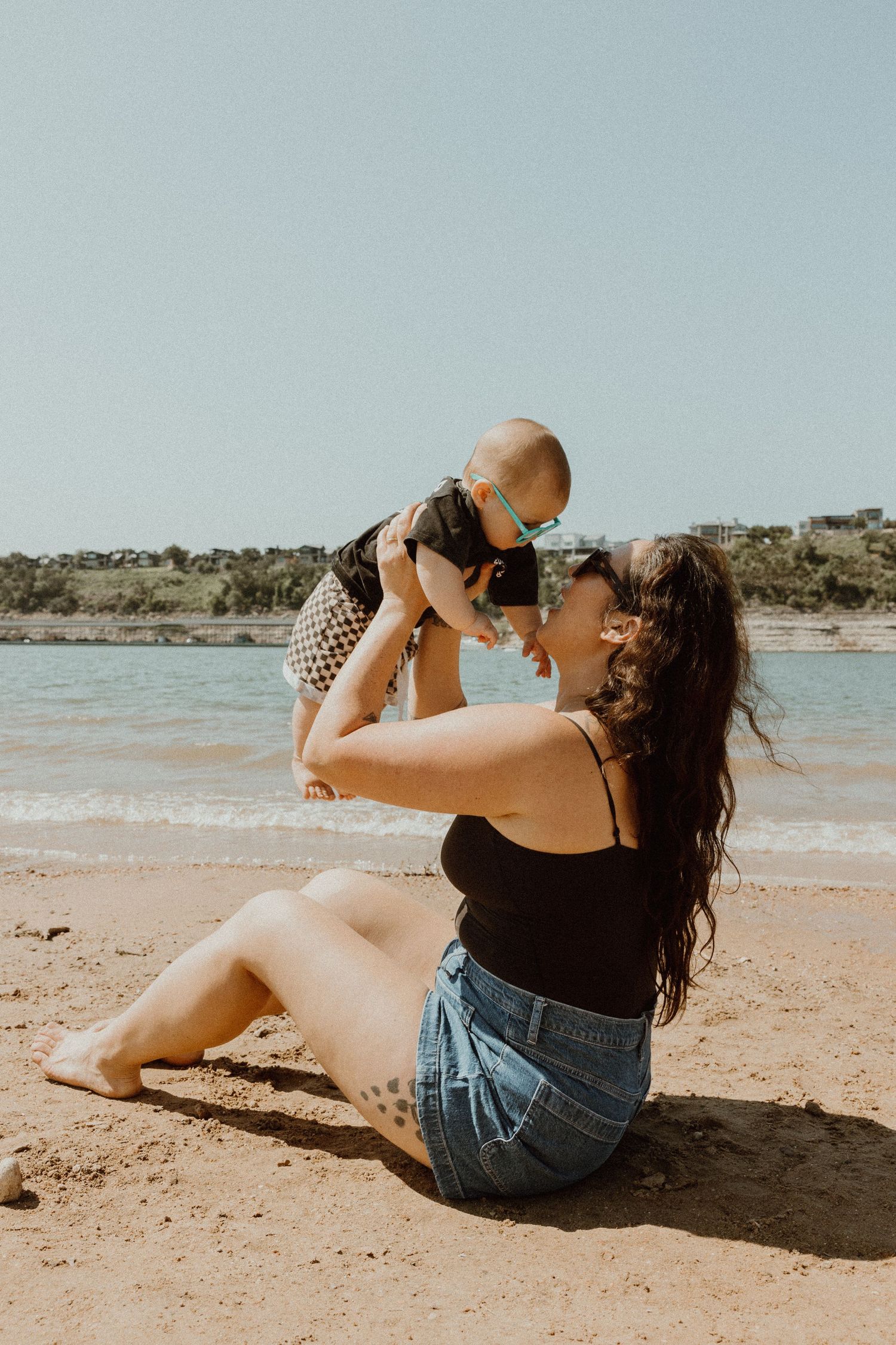 Mother sitting on beach sand lifting smiling baby in the air near ocean waves on sunny day.
