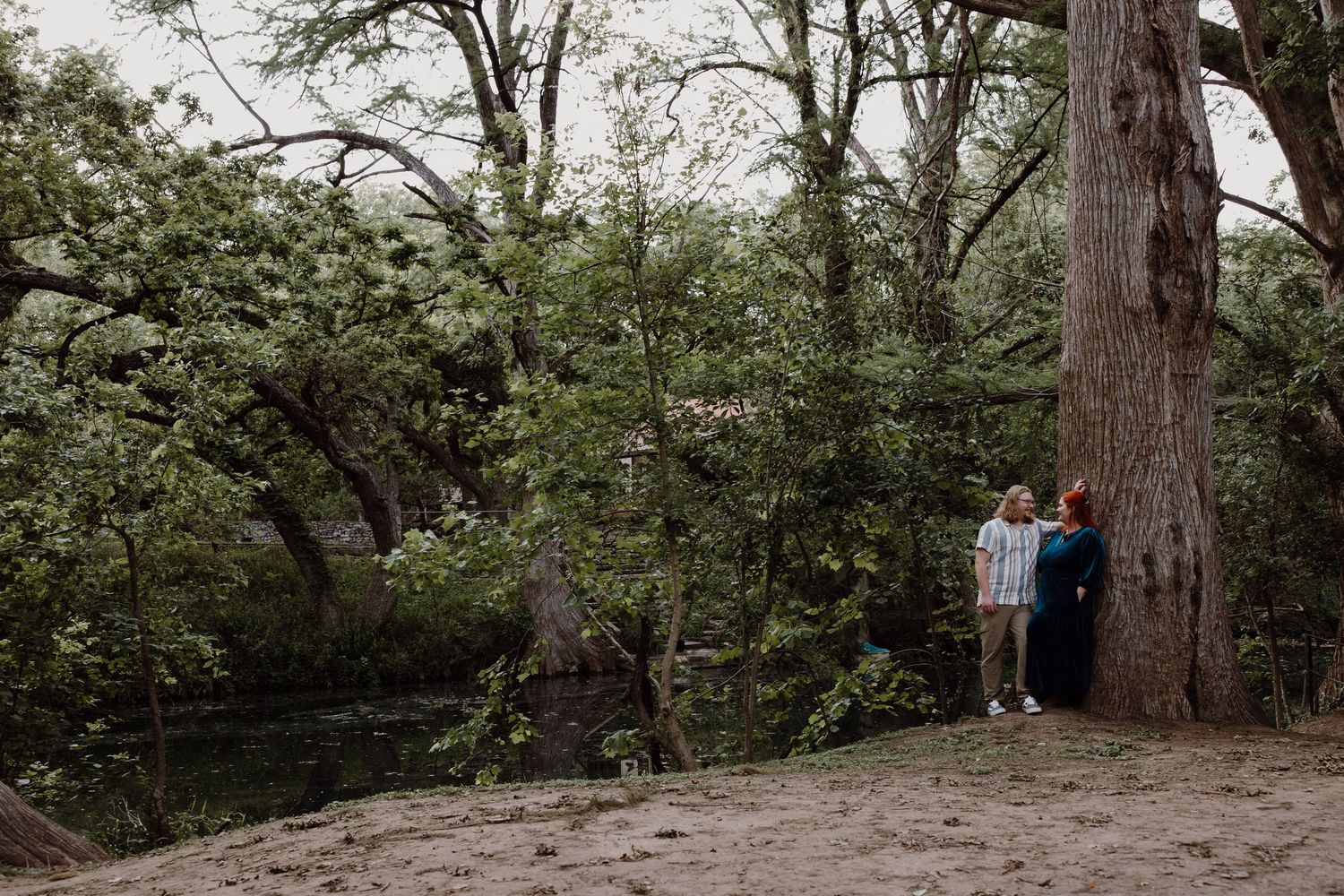 A couple stands together near a large tree by a creek in a wooded outdoor setting on an overcast day.