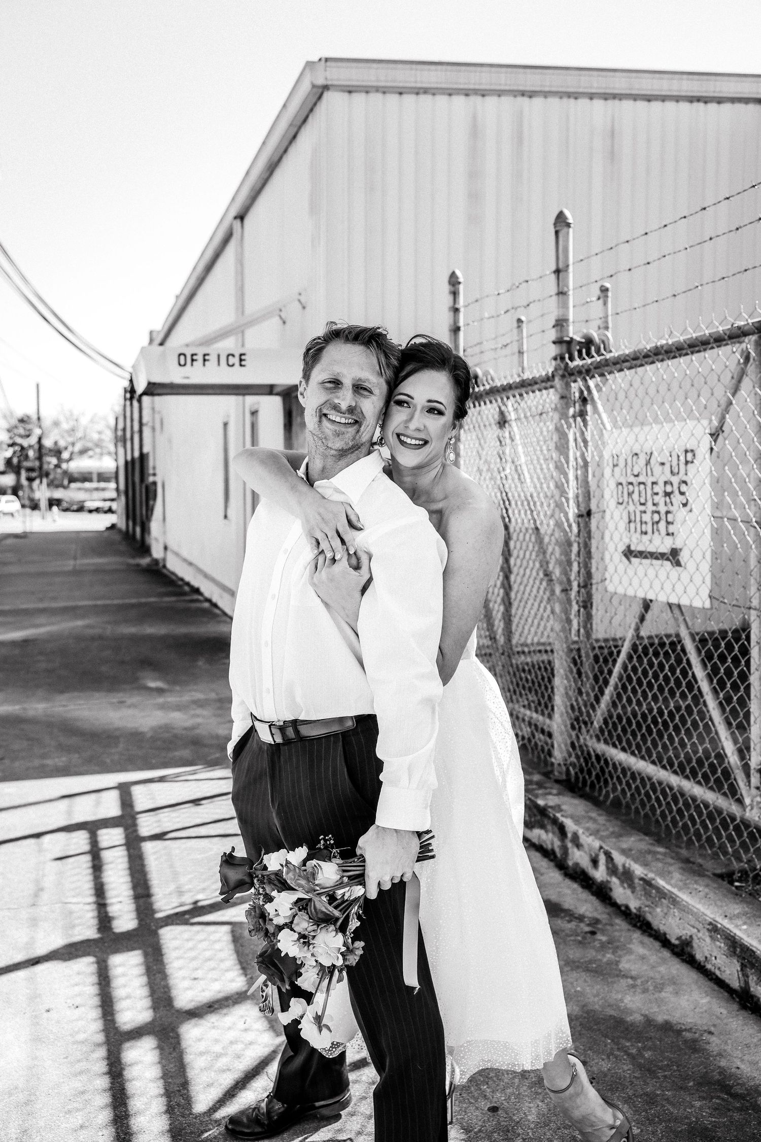 Black and white photo of laughing bride and groom embracing outside an industrial office building.