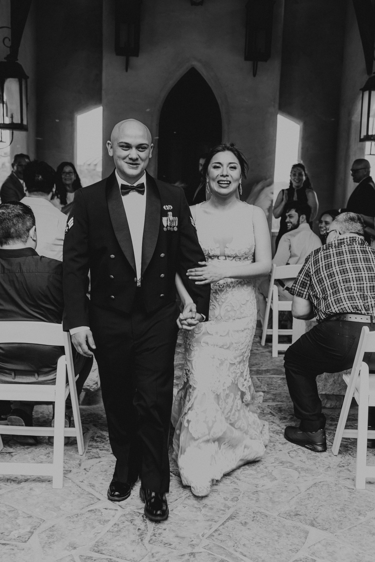 Black and white photo of newlyweds walking down aisle after ceremony, groom in military tuxedo, bride in lace gown.