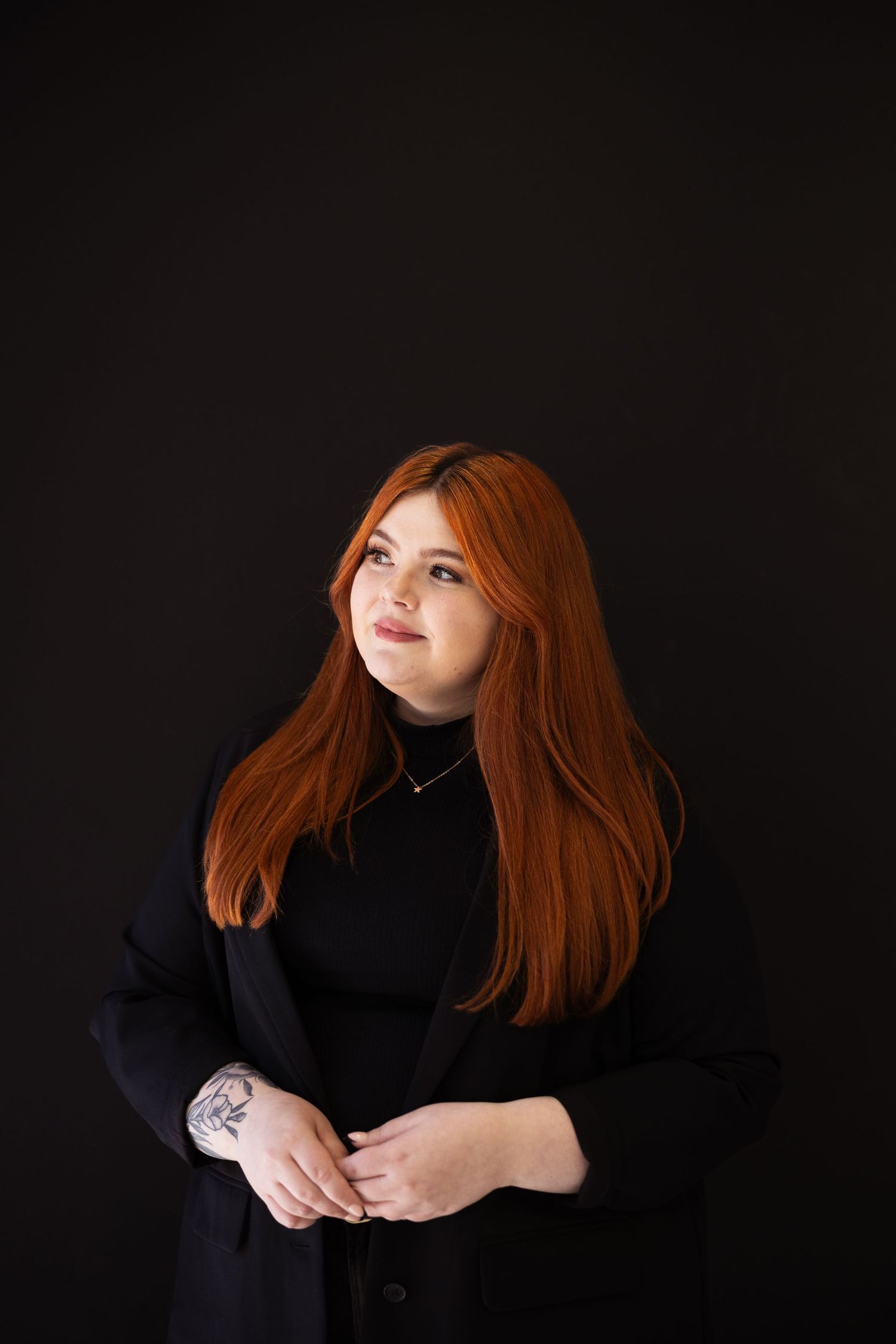 Confident redheaded woman with tattoos in black outfit posing against dark background.