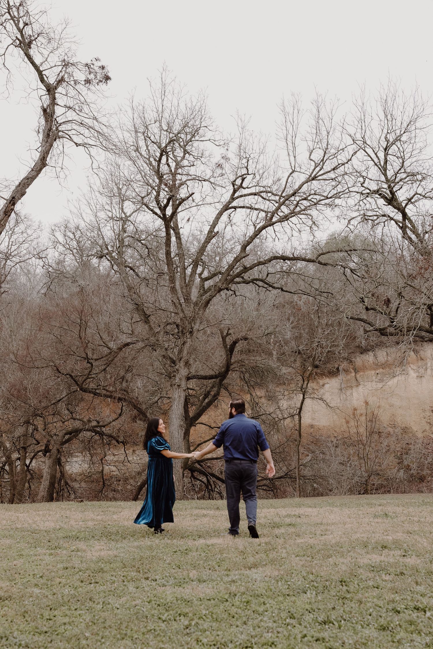 Couple holding hands walking toward a large bare tree in an open field during winter.