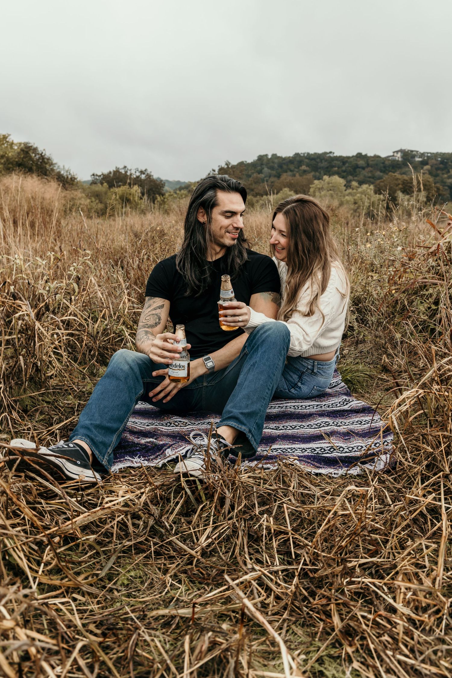 Young couple sitting on a blanket in a dry grass field, clinking beer bottles and smiling.