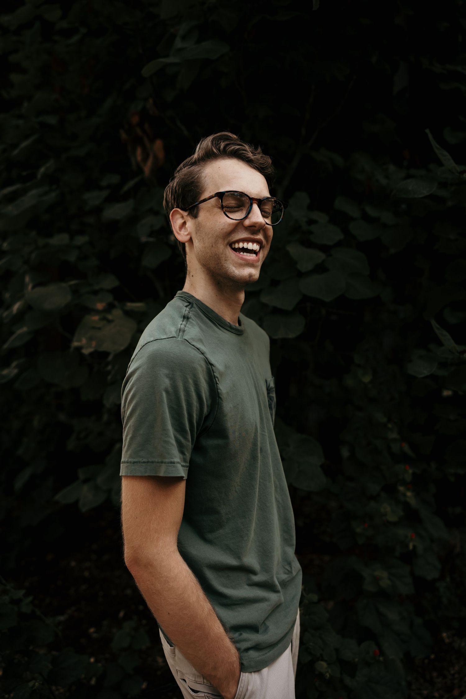 Smiling young man wearing glasses and a green t-shirt stands in front of lush dark foliage.