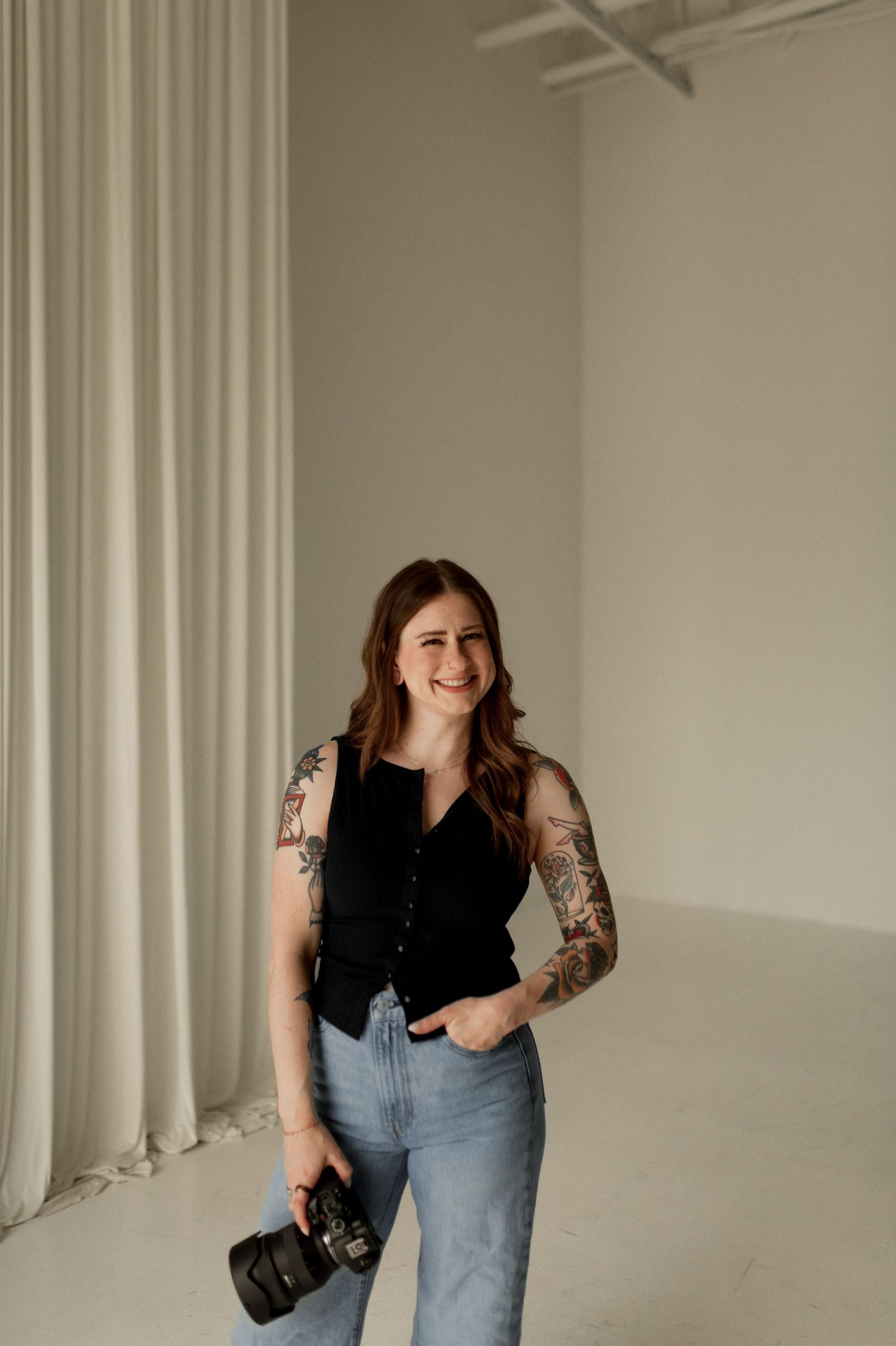 Smiling tattooed female photographer in black top and jeans holding camera in bright studio.