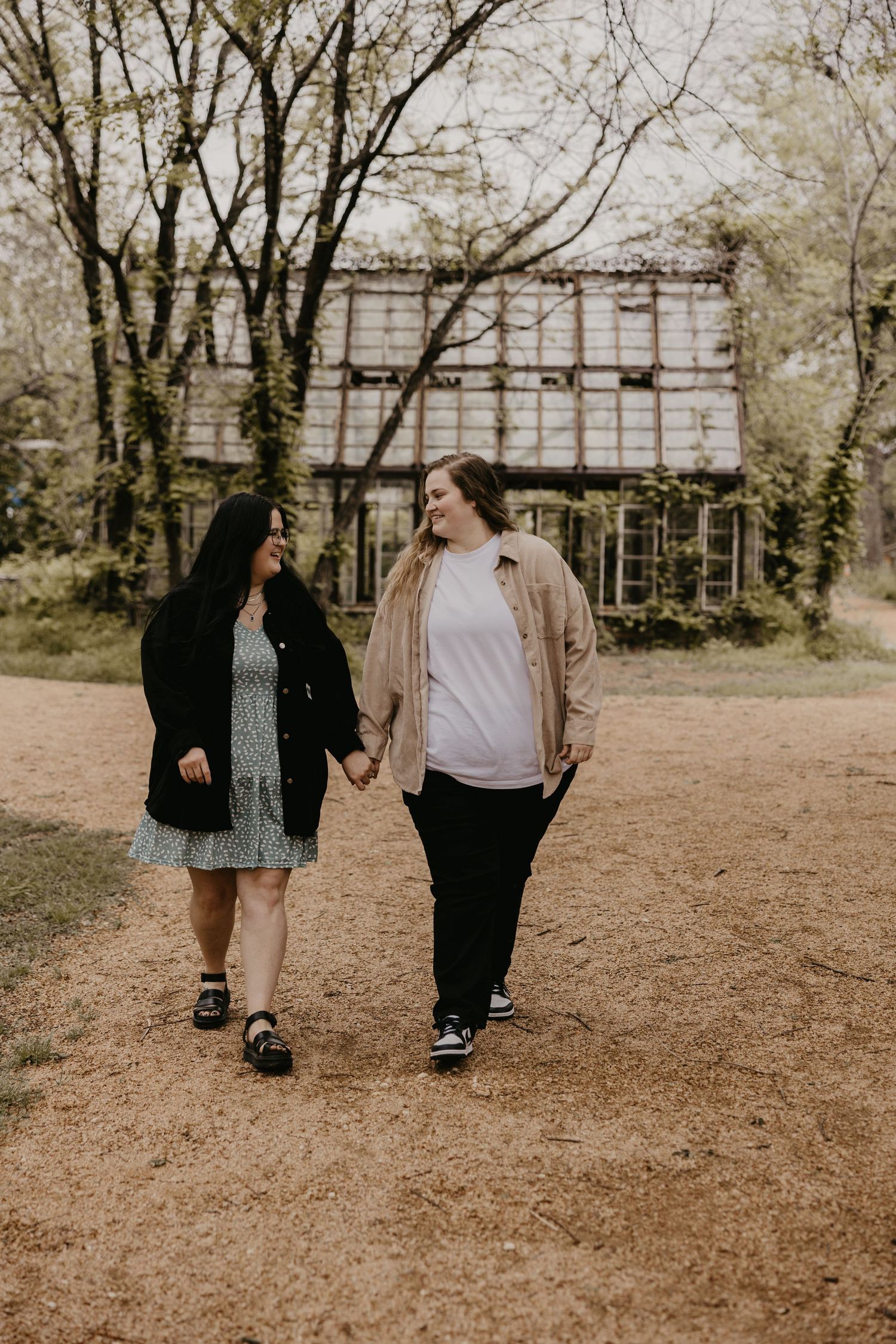 Two women holding hands and smiling while walking near a rustic building outdoors.