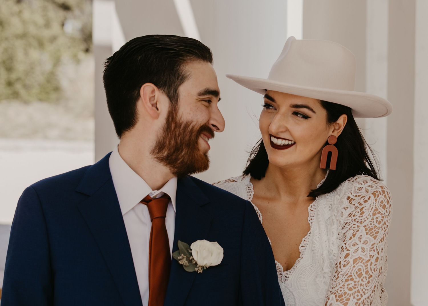 Close-up of smiling bride in white cowboy hat and groom with beard in navy suit on their wedding day.
