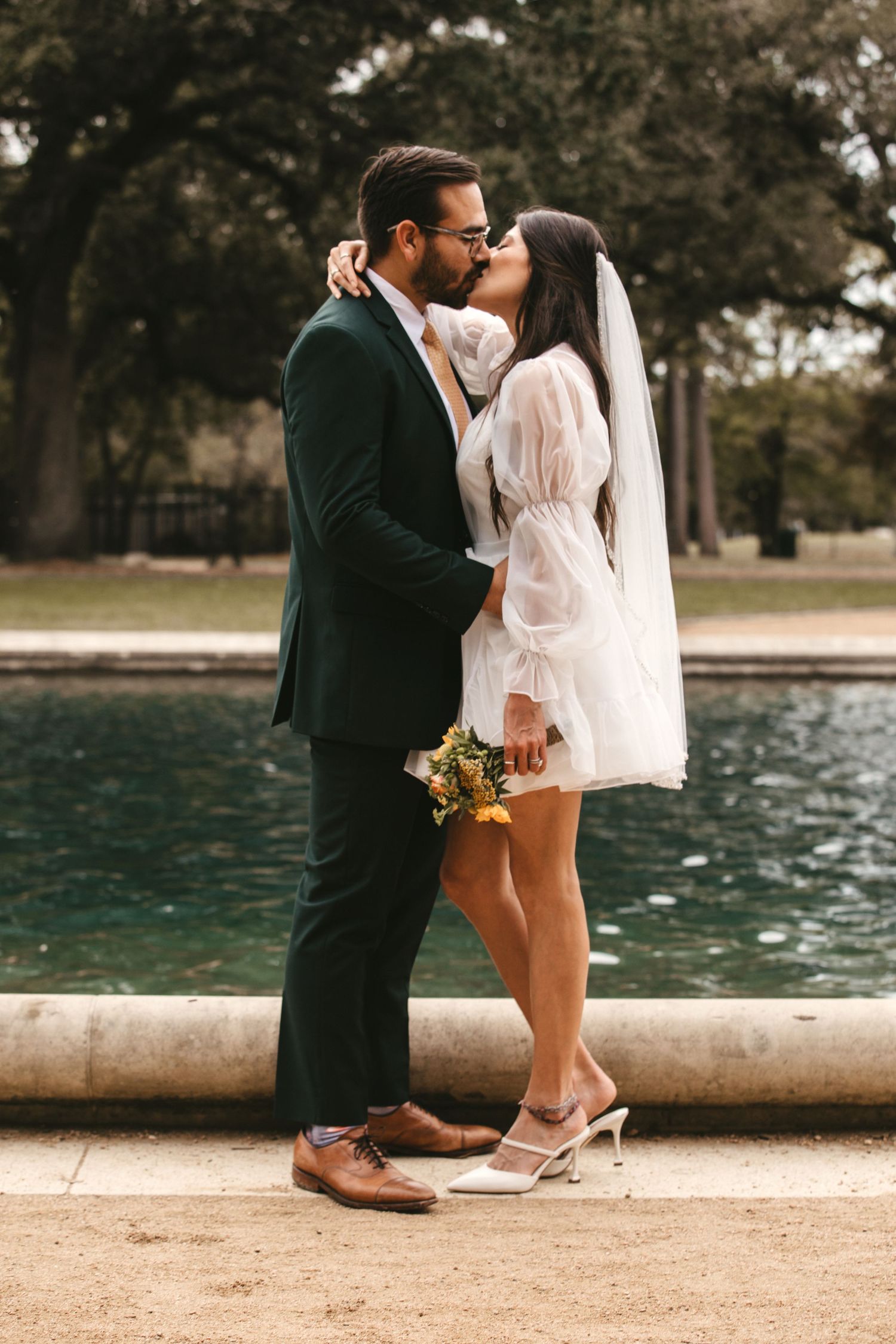 Bride and groom kiss beside a fountain holding a yellow bouquet in outdoor wedding photo.