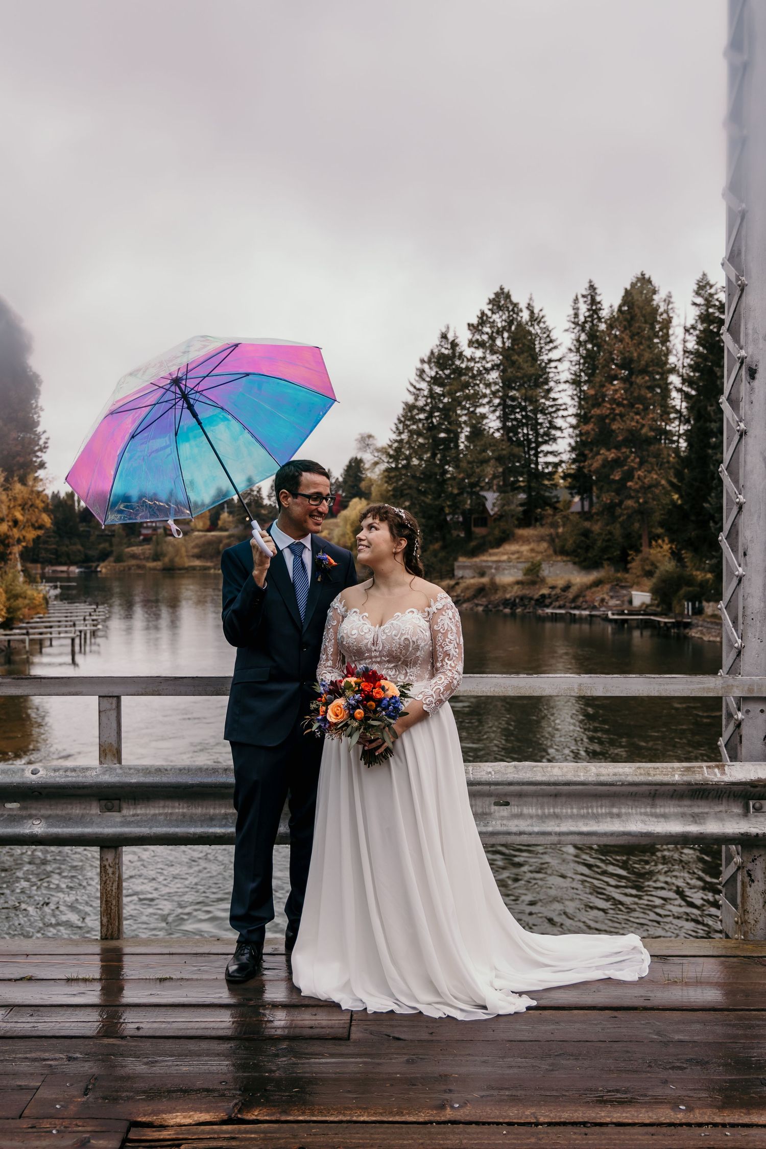 Bride and groom share a joyful moment under a colorful rainbow umbrella on a rainy wedding day by a lake.