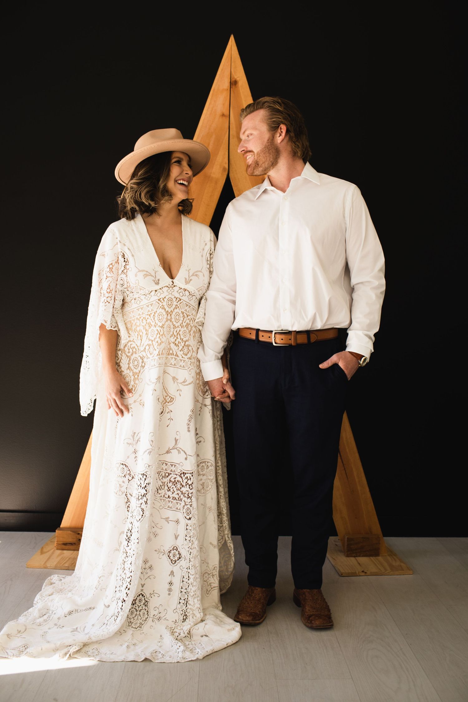 Boho bride in lace dress and hat holds hands with groom in white shirt before geometric wood backdrop.