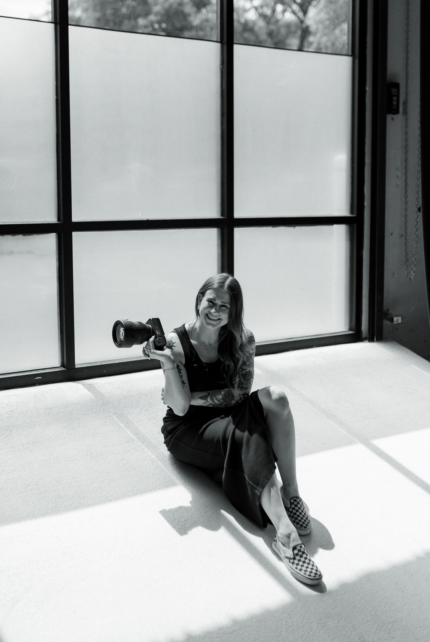 Smiling tattooed woman holding camera sitting on floor by large window in black white.