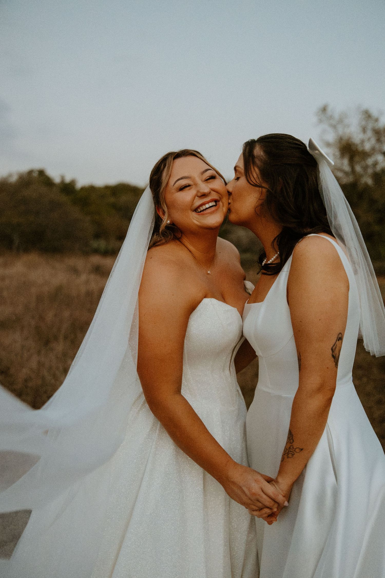 Two brides in white gowns and veils share a tender kiss in a golden field at sunset.