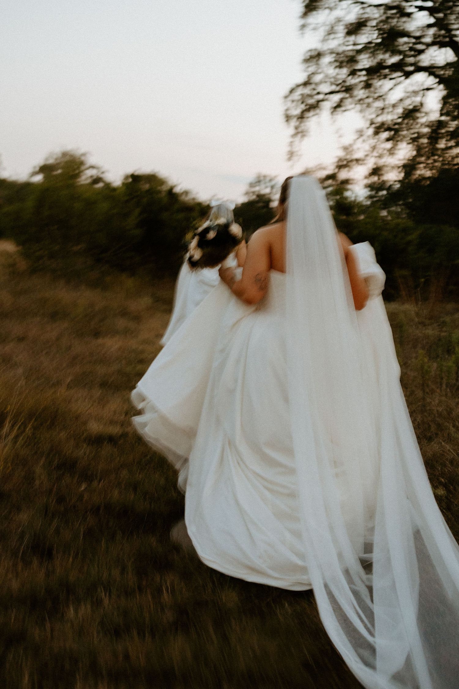 Two brides in white gowns and veils walking through golden field at sunset in motion blur photo.
