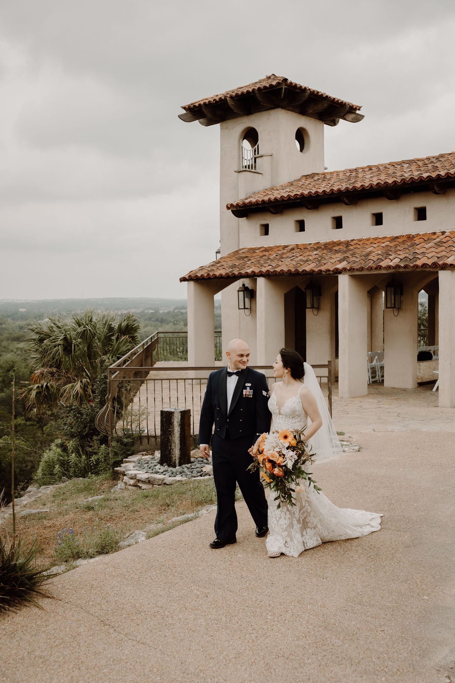 Military groom and lace-gowned bride walk hand in hand outside a Spanish-style wedding venue with panoramic views.